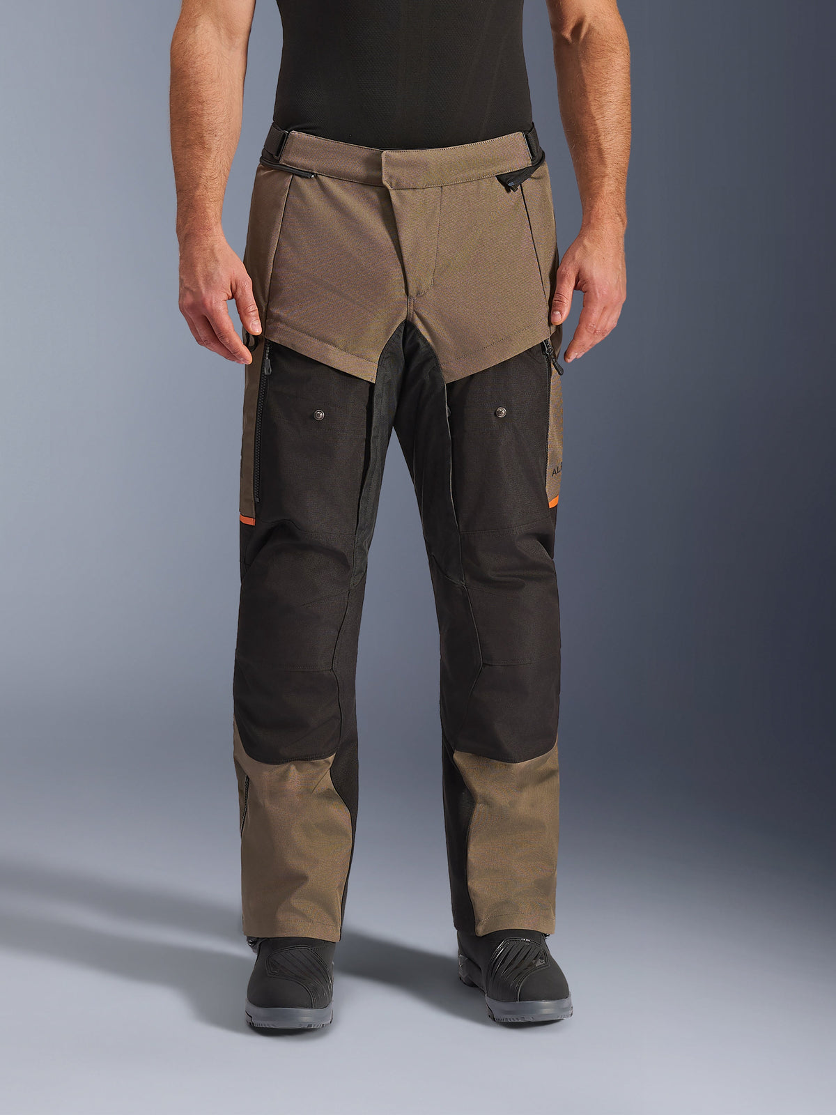 Pantalones Alpinestars Cusco Drystar®, pantalones de motociclismo adventure touring, color nogal y negro con detalles naranja, vista frontal en modelo, con construcción textil técnica, bolsillos cargo y cremalleras de ventilación, usados con botas de motociclista negras