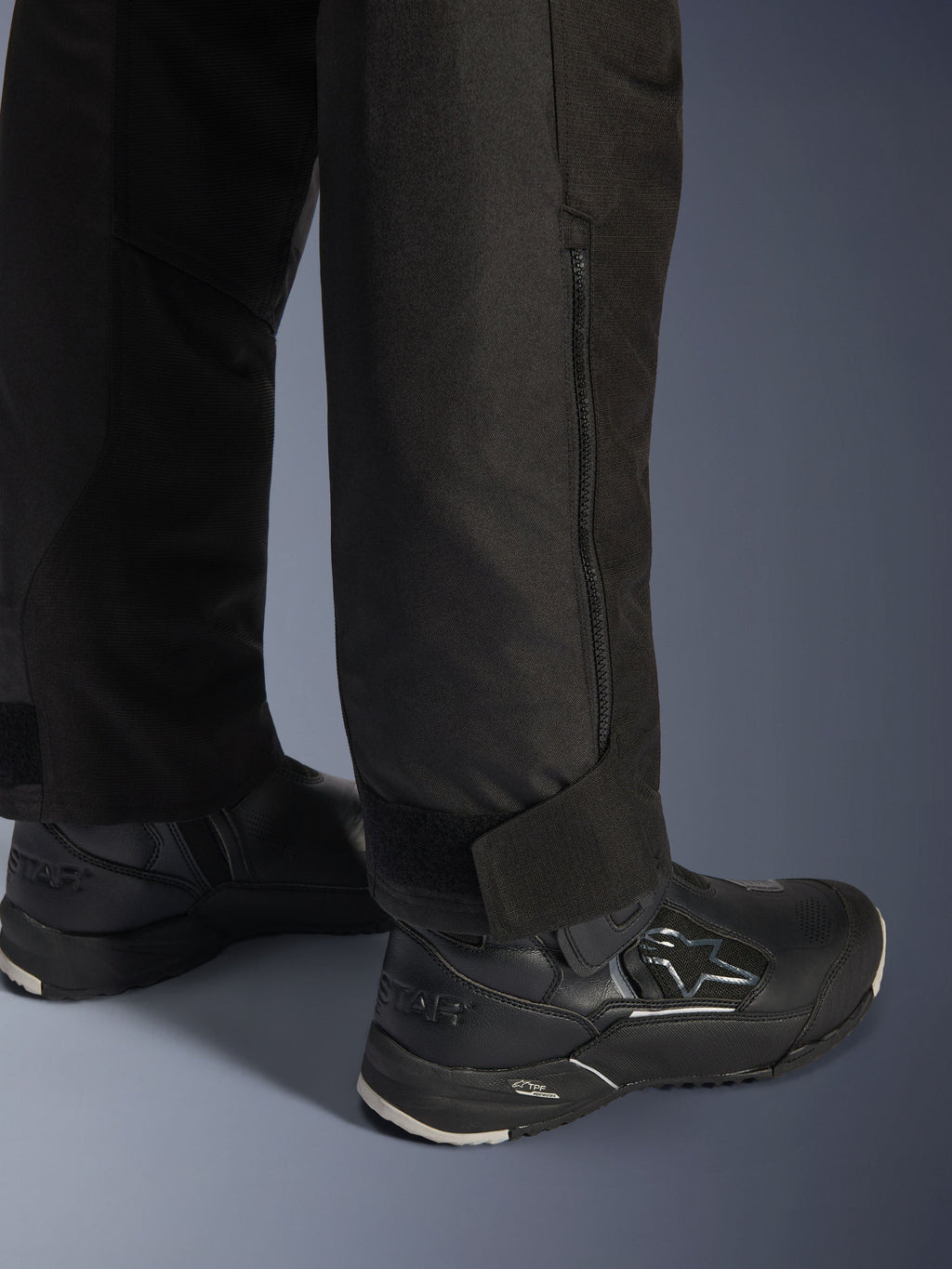 Pantalones Alpinestars Stella Andes V4 Drystar®, negros, vista en primer plano de la parte inferior de la pierna y el bajo, con cremallera lateral y correa de tobillo ajustable de velcro, usados con botas de conducción Alpinestars negras