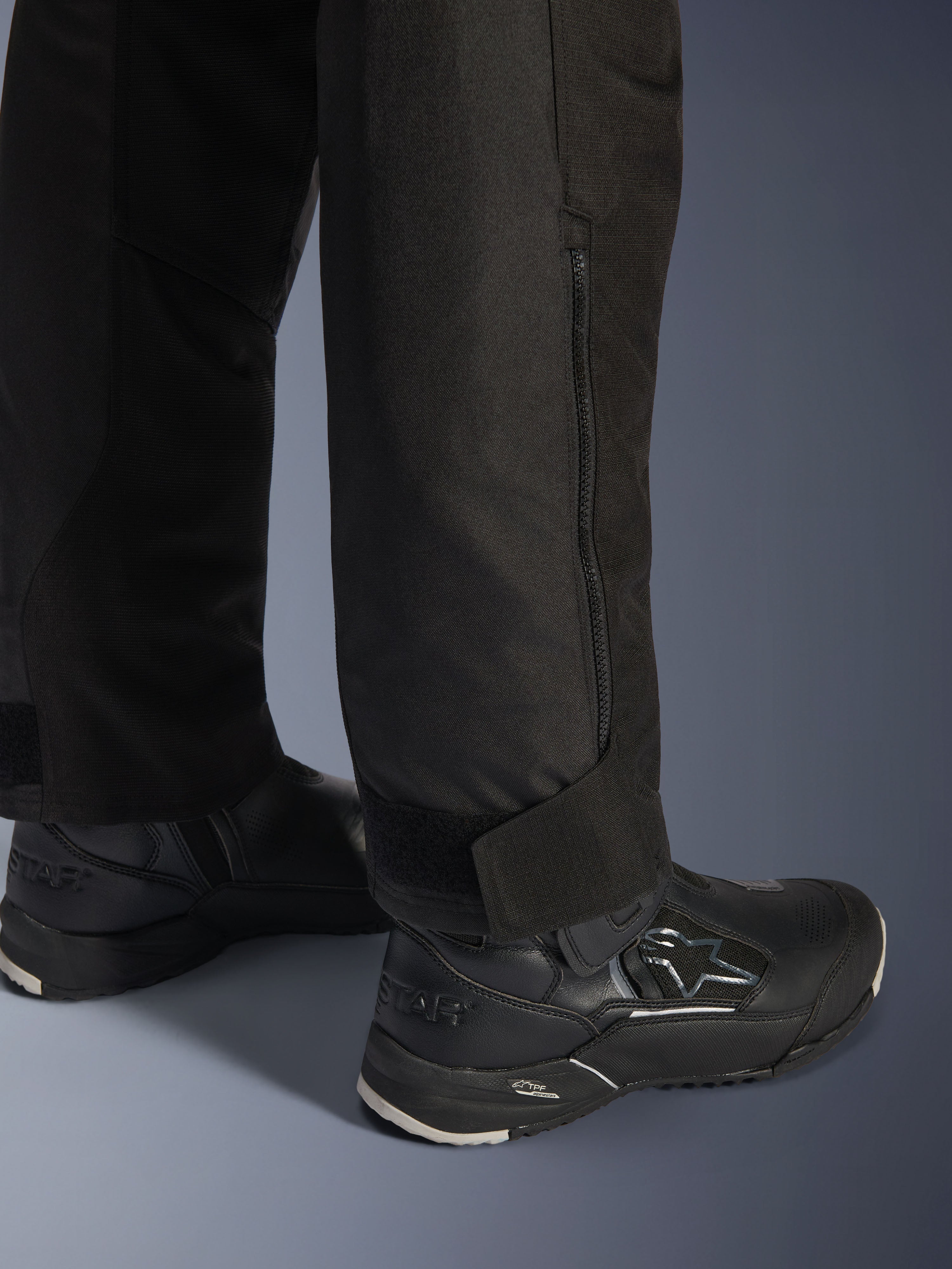 Pantalones Alpinestars Stella Andes V4 Drystar®, negros, vista en primer plano de la parte inferior de la pierna y el bajo, con cremallera lateral y correa de tobillo ajustable de velcro, usados con botas de conducción Alpinestars negras