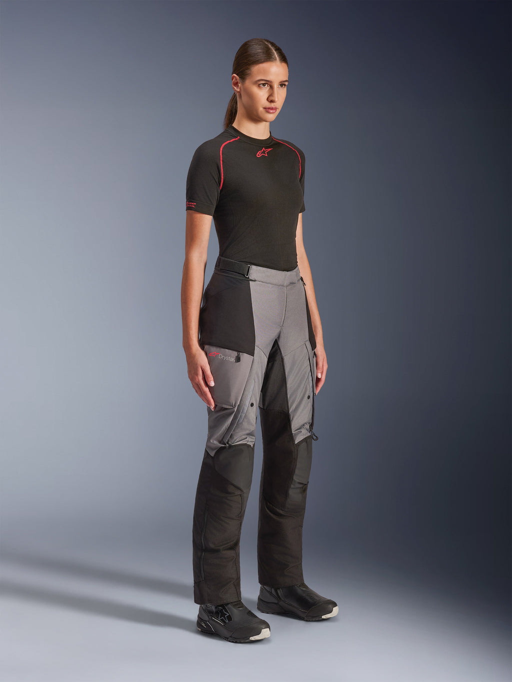 Pantalones Alpinestars Stella Andes V4 Drystar®, pantalones de moto de aventura para mujer, negro y gris oscuro, lucidos por una modelo con una capa base Alpinestars negra y botas negras, con paneles reforzados, logotipo Drystar rojo y bolsillos cargo.
