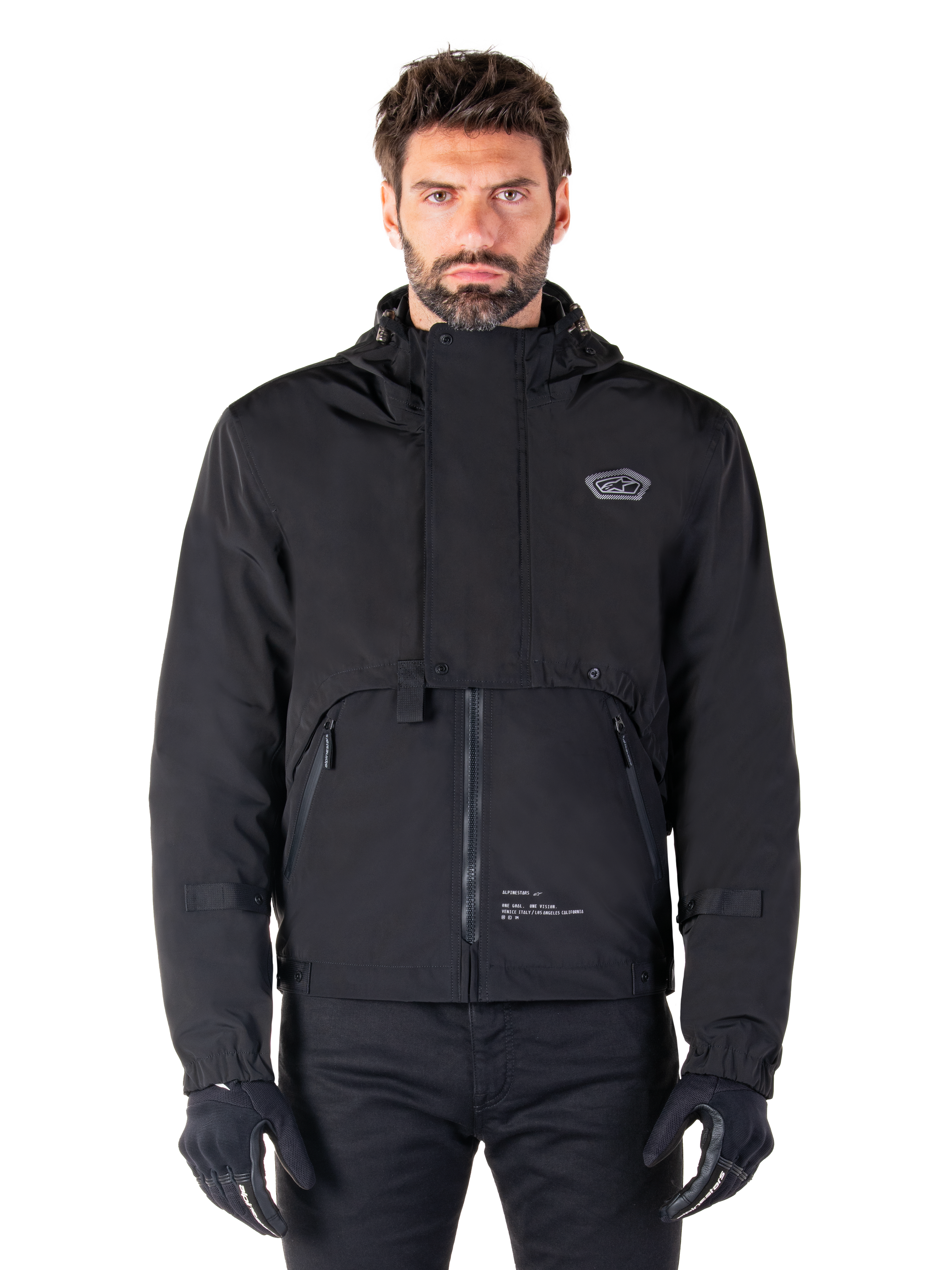 Alpinestars Mootant Waterproof Parka, chaqueta de moto negra con capucha con un diseño técnico modular, usada por un modelo masculino con guantes negros, con una solapa frontal única y bolsillos laterales con cremallera, estilo de conducción urbano y touring.