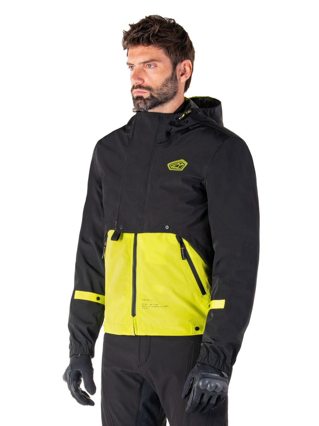 Alpinestars Mootant Waterproof Parka en Lime Punch Black, chaqueta modular de moto usada por un hombre con guantes y pantalones de montar negros, con una parte superior y mangas negras y un panel inferior verde lima vibrante, diseño con capucha de estética utilitaria inspirada en lo militar.
