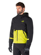 Alpinestars Mootant Waterproof Parka en Lime Punch Black, chaqueta modular de moto usada por un hombre con guantes y pantalones de montar negros, con una parte superior y mangas negras y un panel inferior verde lima vibrante, diseño con capucha de estética utilitaria inspirada en lo militar.