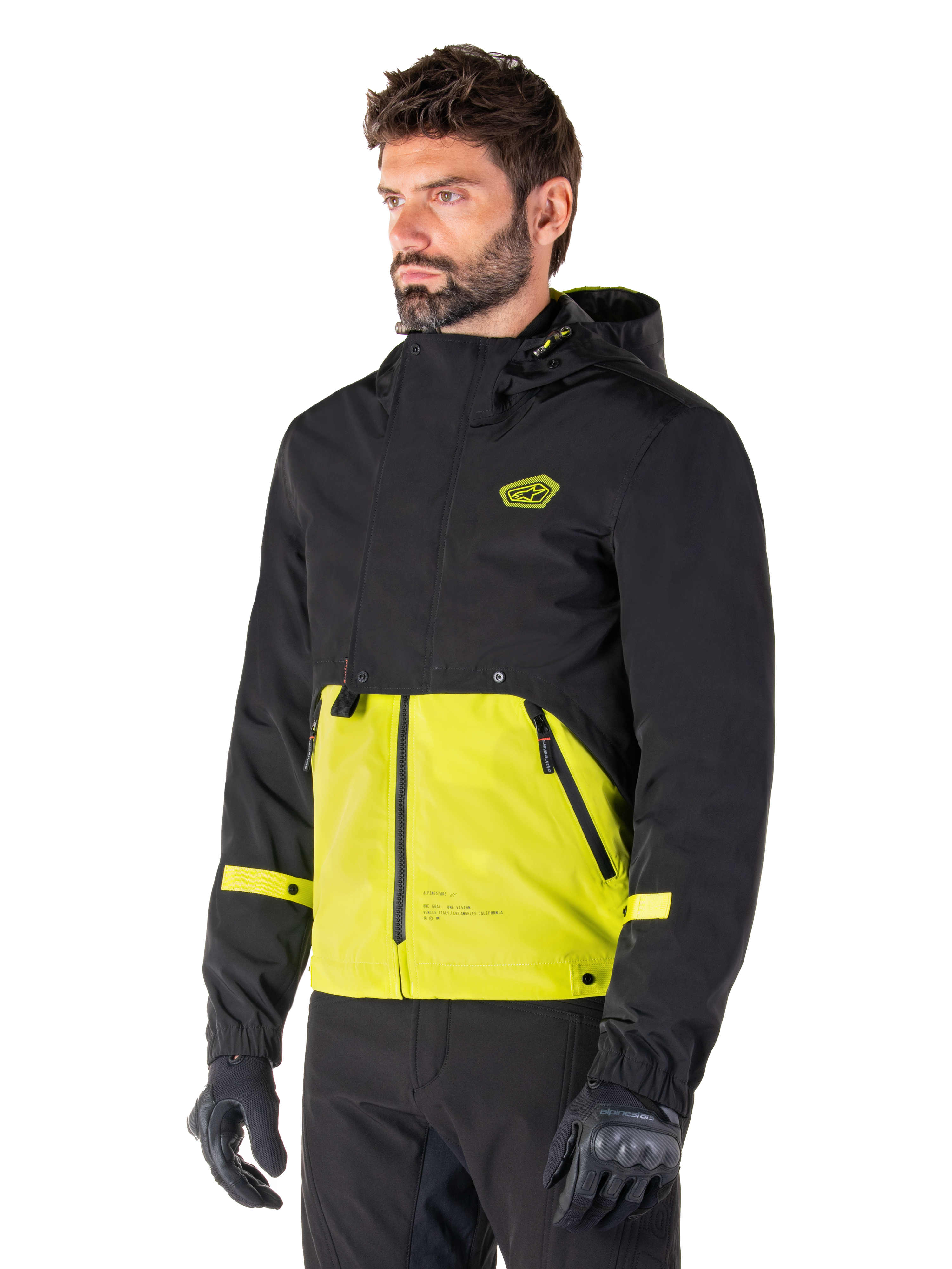 Alpinestars Mootant Waterproof Parka en Lime Punch Black, chaqueta modular de moto usada por un hombre con guantes y pantalones de montar negros, con una parte superior y mangas negras y un panel inferior verde lima vibrante, diseño con capucha de estética utilitaria inspirada en lo militar.