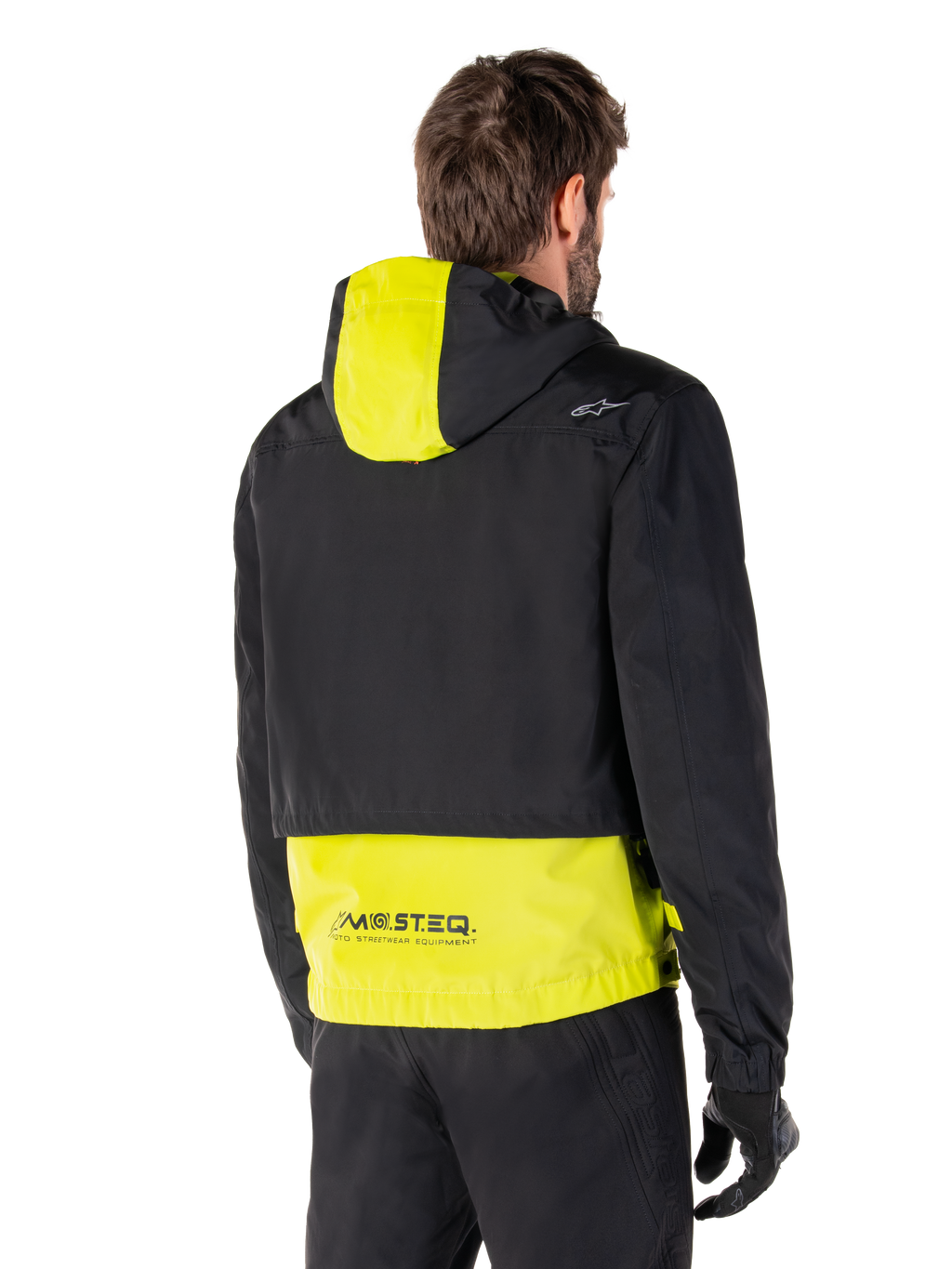 Parka impermeable Alpinestars Mootant, vista trasera, verde lima y negro, con la parte superior del cuerpo en negro y el panel de la capucha y la parte inferior de la espalda en amarillo lima, marca MO.ST.EQ. en el dobladillo, combinada con pantalones y guantes de moto negros, chaqueta de moto urbana contemporánea