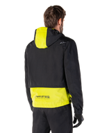 Parka impermeable Alpinestars Mootant, vista trasera, verde lima y negro, con la parte superior del cuerpo en negro y el panel de la capucha y la parte inferior de la espalda en amarillo lima, marca MO.ST.EQ. en el dobladillo, combinada con pantalones y guantes de moto negros, chaqueta de moto urbana contemporánea