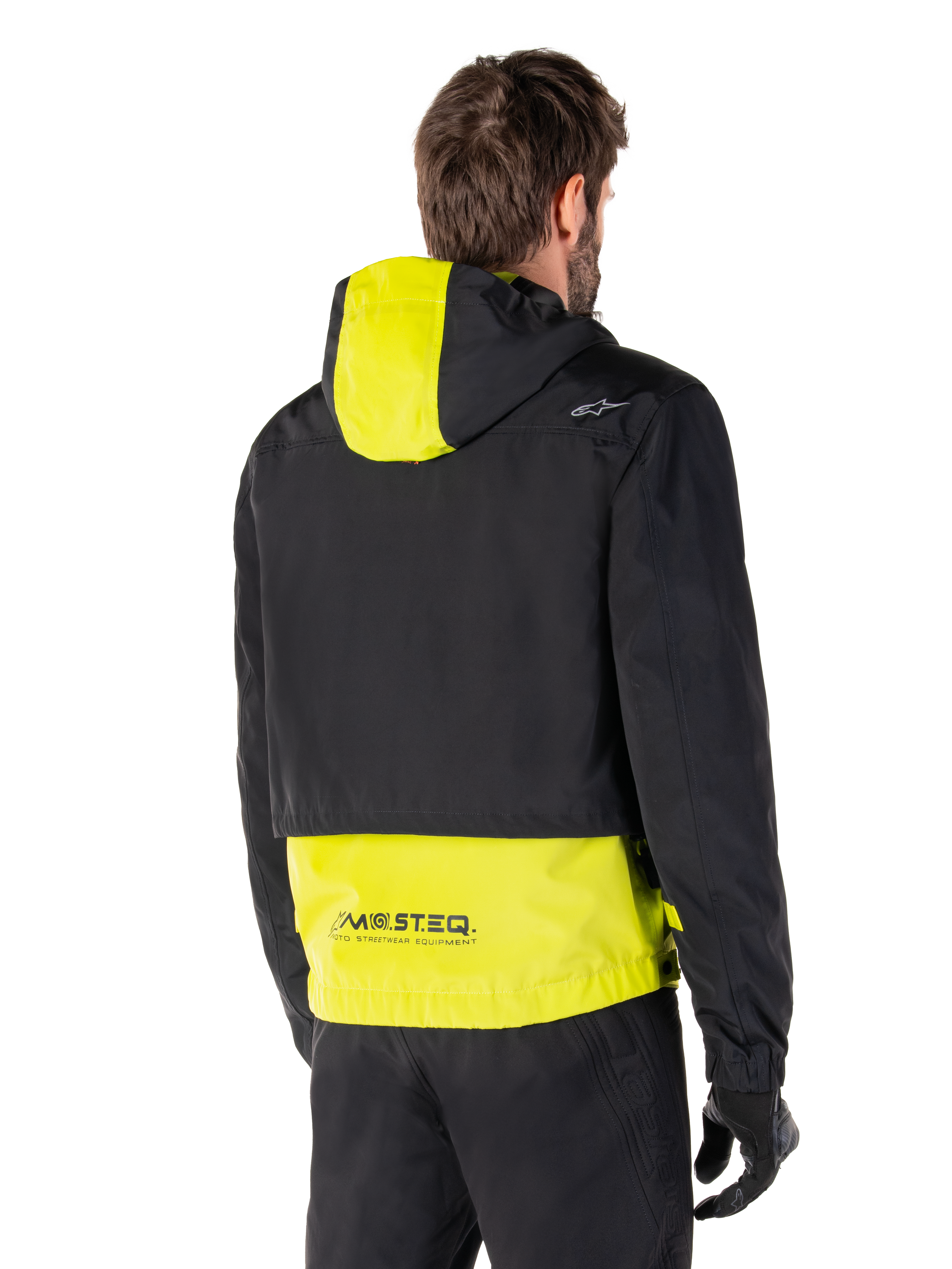 Parka impermeable Alpinestars Mootant, vista trasera, verde lima y negro, con la parte superior del cuerpo en negro y el panel de la capucha y la parte inferior de la espalda en amarillo lima, marca MO.ST.EQ. en el dobladillo, combinada con pantalones y guantes de moto negros, chaqueta de moto urbana contemporánea