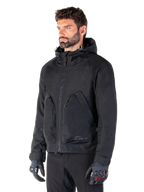 Man wearing Alpinestars Morush Waterproof Parka, Black urban motorcycle jacket, streetwear-inspired design with hood and large zippered front pockets, paired with grey and black motorcycle gloves, contemporary minimalist style