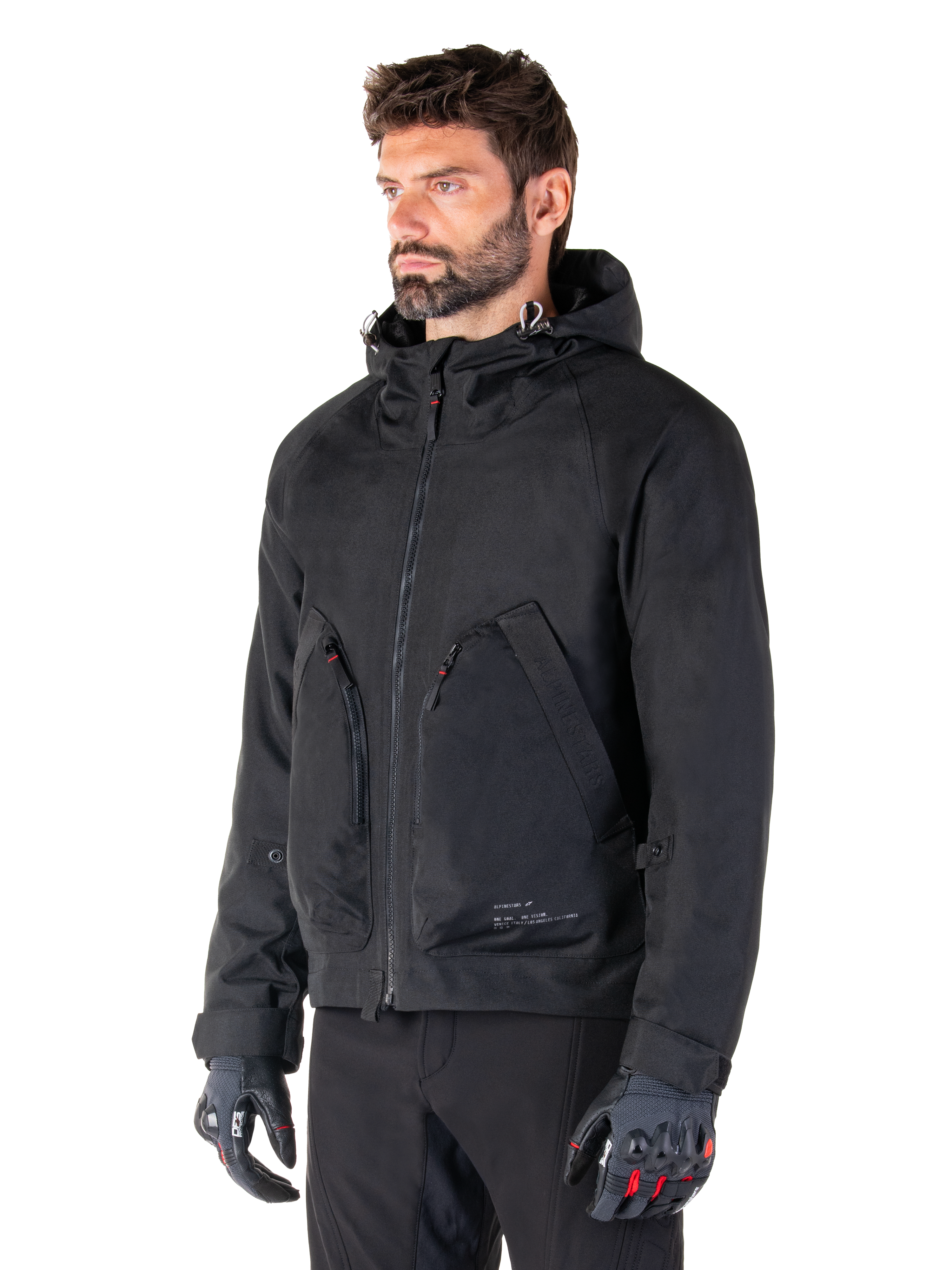 Man wearing Alpinestars Morush Waterproof Parka, Black urban motorcycle jacket, streetwear-inspired design with hood and large zippered front pockets, paired with grey and black motorcycle gloves, contemporary minimalist style