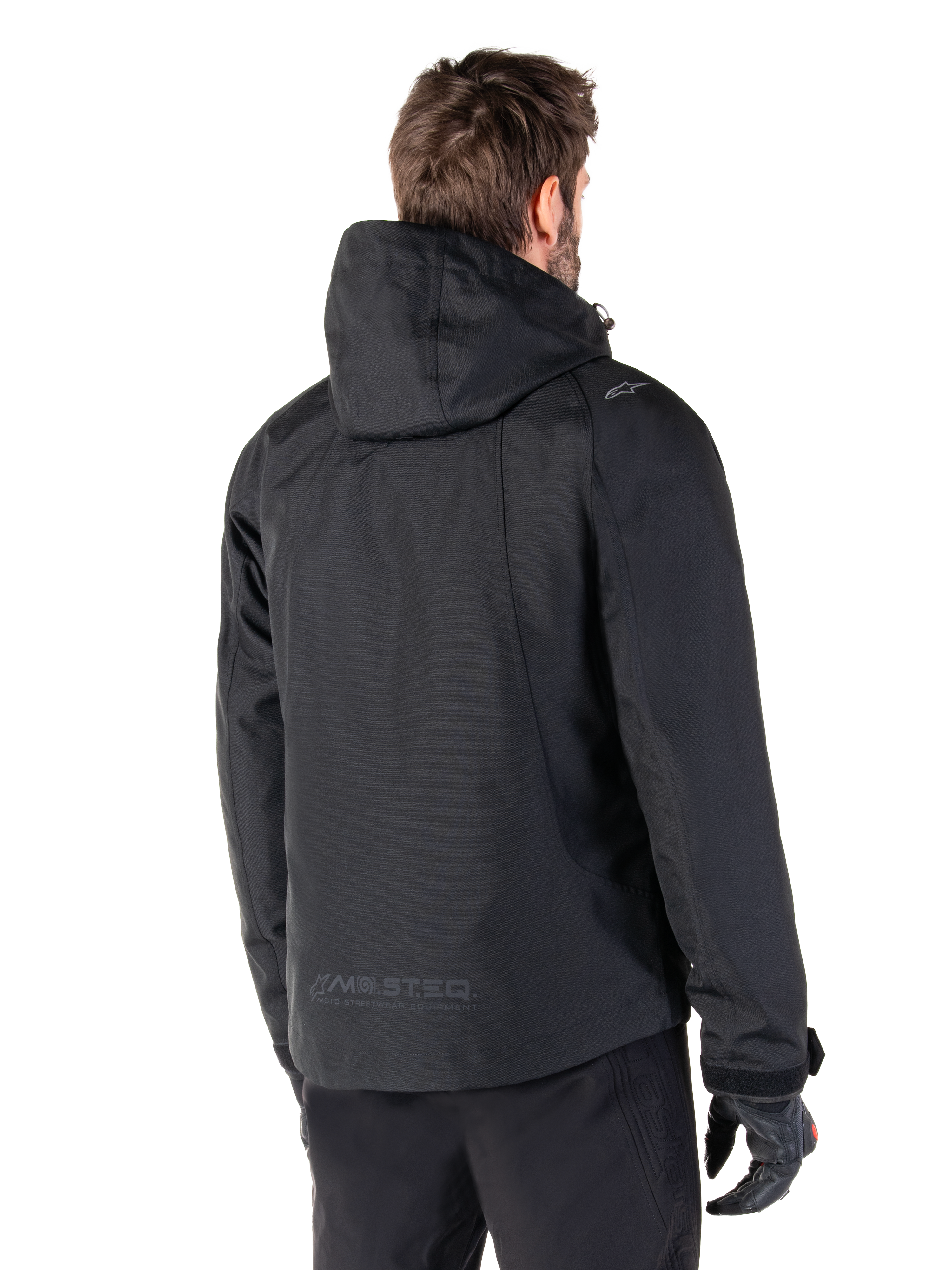Alpinestars Morush Waterproof Parka, Black, rear view of a man wearing the hooded motorcycle jacket, featuring raglan sleeves, an extended rear hem for coverage, MO.ST.EQ. branding on the lower back, and a subtle Alpinestars logo on the shoulder, urban streetwear style