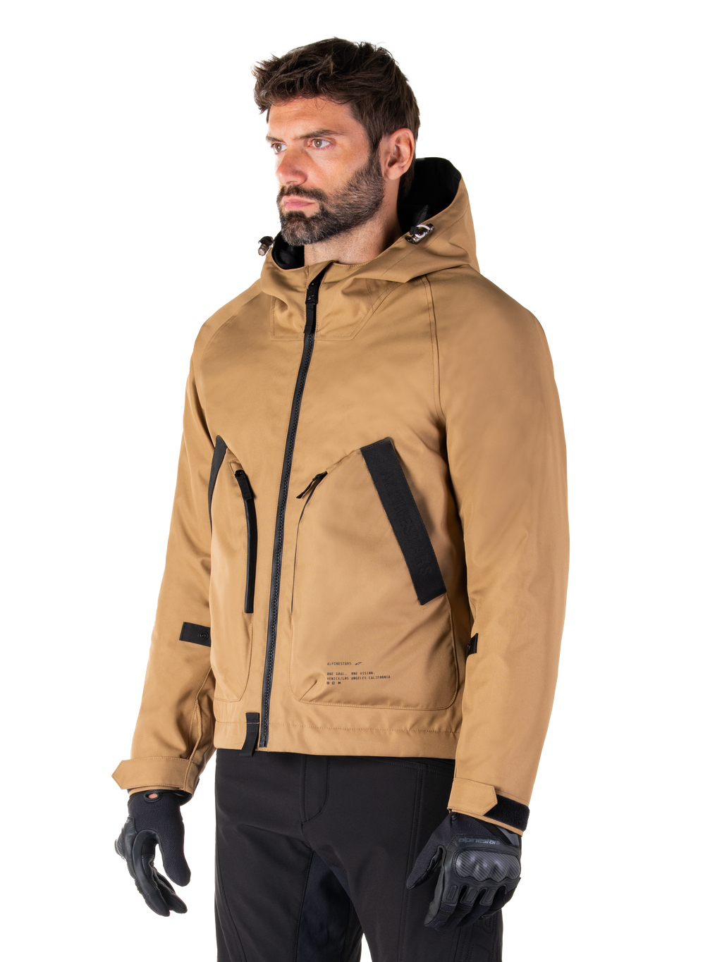Alpinestars Morush Waterproof Parka, marrón Utility, modelo masculino vistiendo la parka con guantes de conducción negros y pantalones negros, diseño minimalista inspirado en la moda urbana con capucha a prueba de tormentas y grandes bolsillos frontales, ropa de motociclismo impermeable