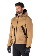 Alpinestars Morush Waterproof Parka, marrón Utility, modelo masculino vistiendo la parka con guantes de conducción negros y pantalones negros, diseño minimalista inspirado en la moda urbana con capucha a prueba de tormentas y grandes bolsillos frontales, ropa de motociclismo impermeable