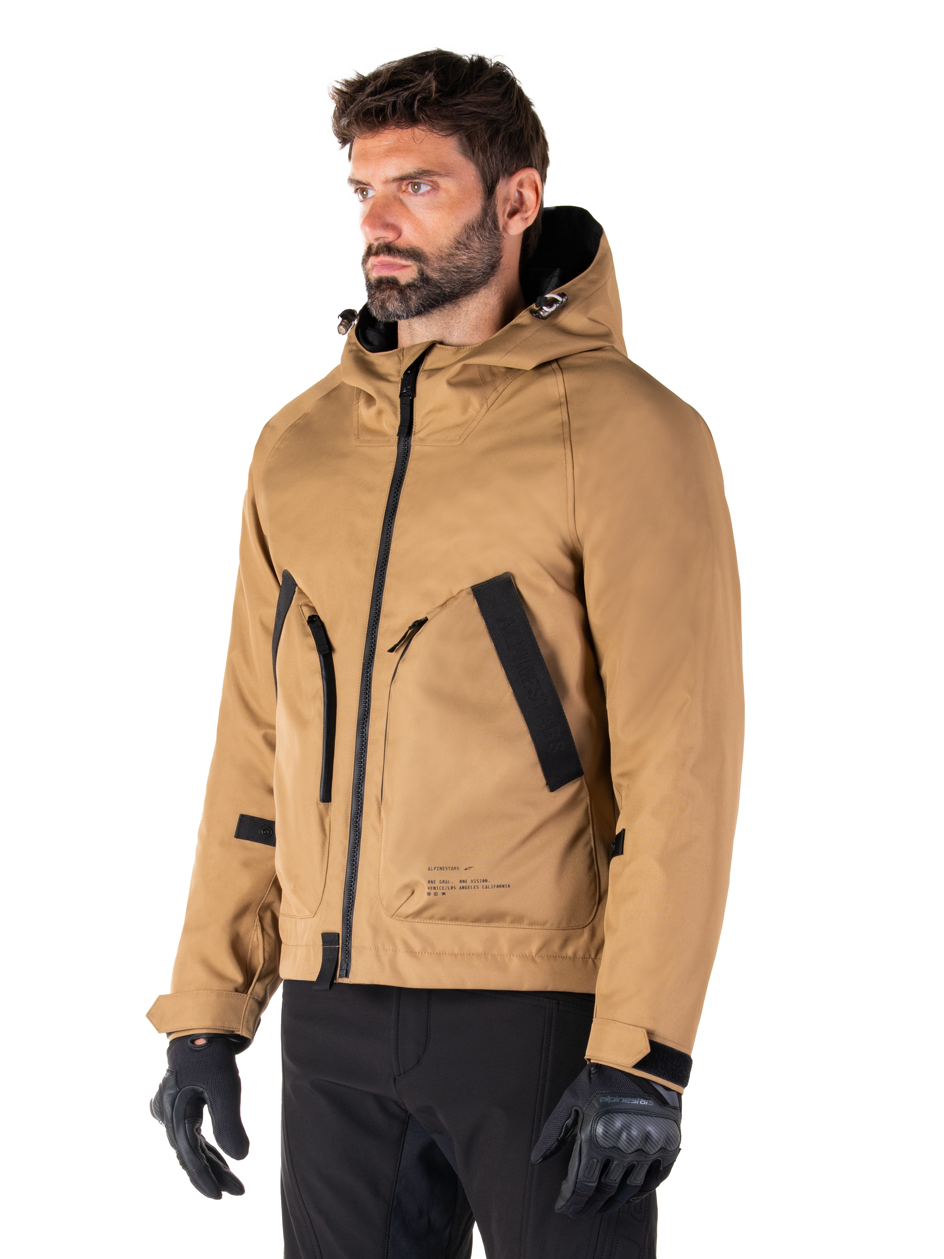 Alpinestars Morush Waterproof Parka, marrón Utility, modelo masculino vistiendo la parka con guantes de conducción negros y pantalones negros, diseño minimalista inspirado en la moda urbana con capucha a prueba de tormentas y grandes bolsillos frontales, ropa de motociclismo impermeable