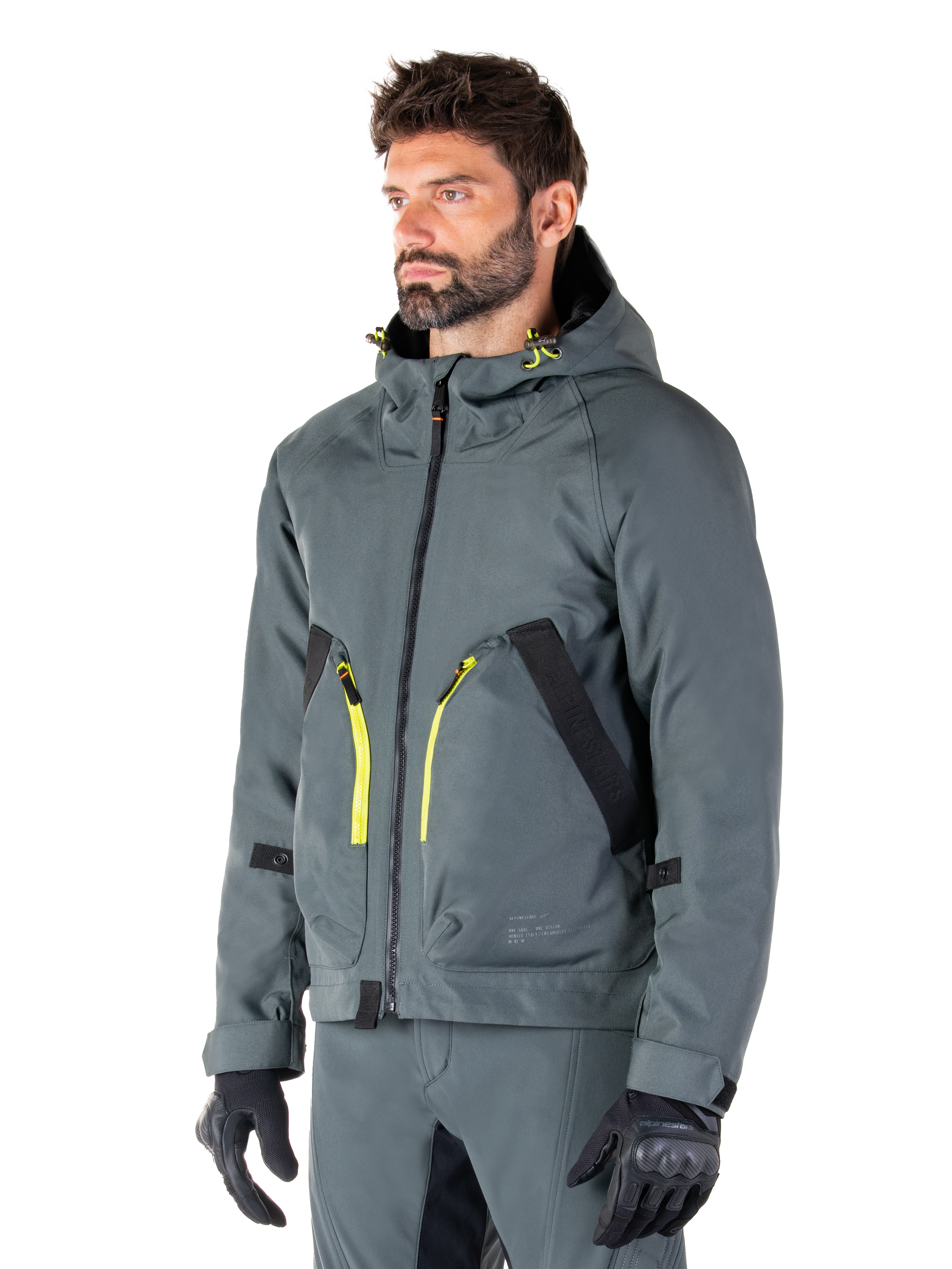 Parka impermeable Alpinestars Morush, chaqueta de moto en Urban Gray, diseño con capucha con detalles de cremalleras en amarillo brillante, detalles de bolsillos en negro, combinada con pantalones de conducción grises a juego y guantes negros, ropa de motociclismo contemporánea de inspiración urbana.