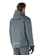 Parka impermeable Alpinestars Morush en Urban Gray, vista trasera, con la marca MO.ST.EQ. en amarillo en la parte inferior de la espalda y el hombro, diseño minimalista con capucha, combinada con pantalones de conducción grises a juego y guantes negros, estilo urbano para motociclismo.