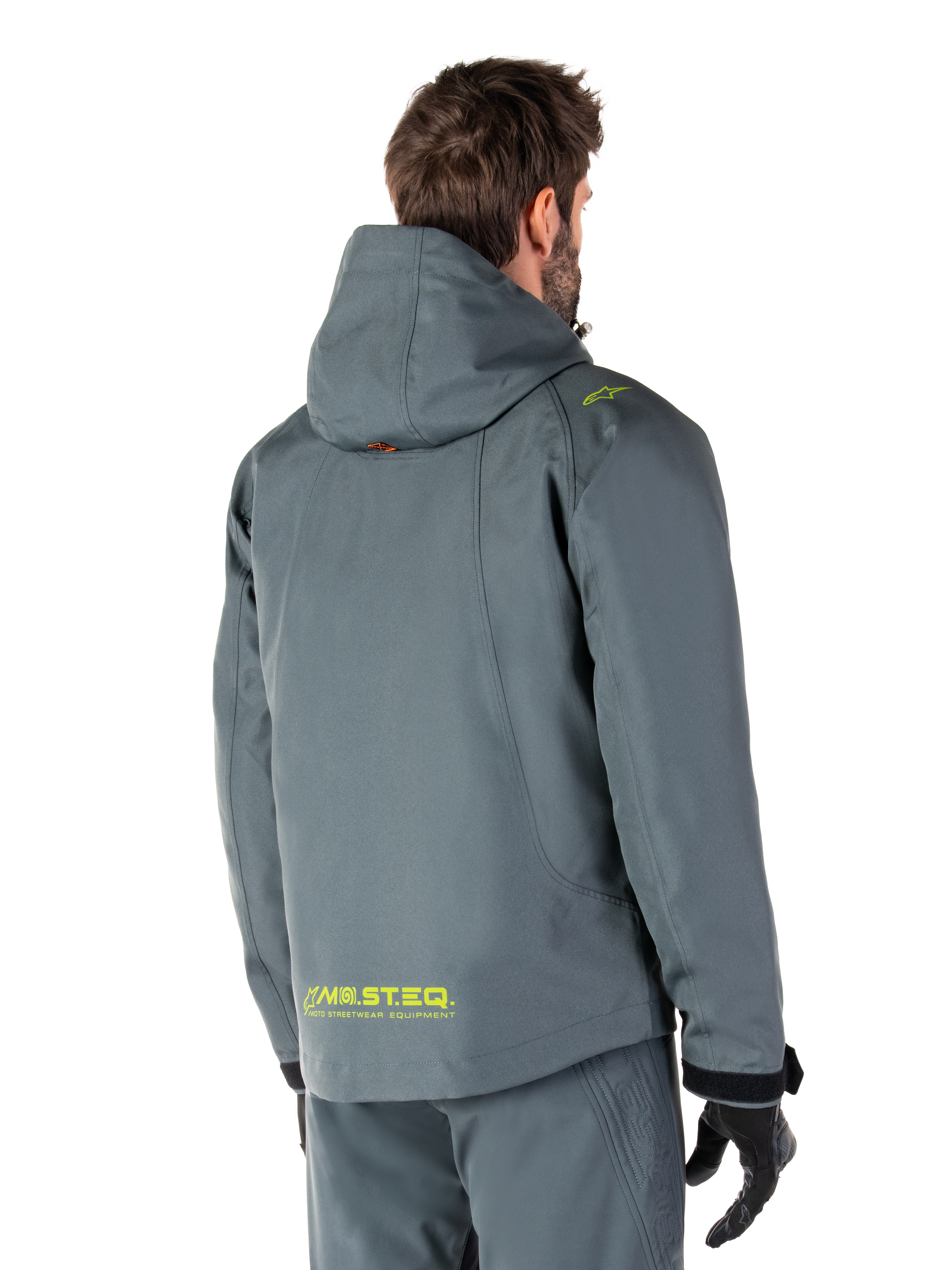 Parka impermeable Alpinestars Morush en Urban Gray, vista trasera, con la marca MO.ST.EQ. en amarillo en la parte inferior de la espalda y el hombro, diseño minimalista con capucha, combinada con pantalones de conducción grises a juego y guantes negros, estilo urbano para motociclismo.