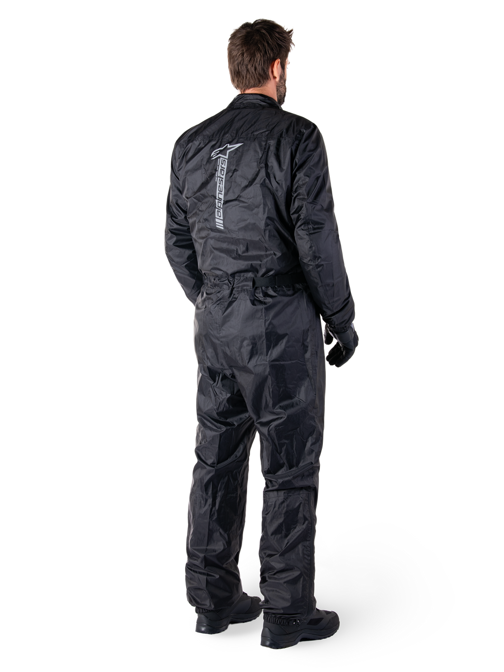Alpinestars Hurricane Rain V2 Suit, mono de lluvia impermeable para motocicleta de una pieza en color negro, vista trasera con gran logotipo reflectante vertical, tejido ligero resistente al viento, usado con guantes y botas negras
