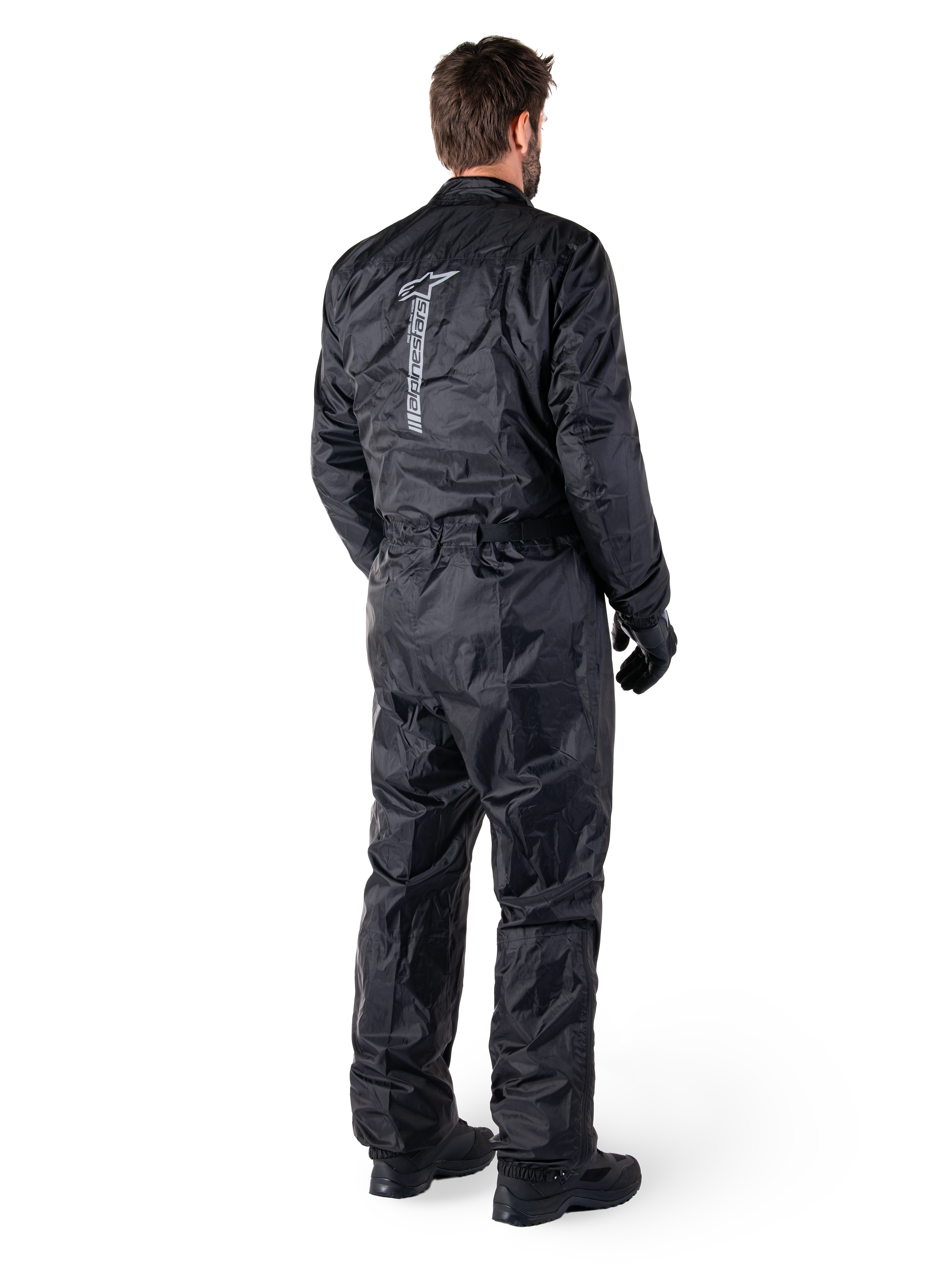 Alpinestars Hurricane Rain V2 Suit, mono de lluvia impermeable para motocicleta de una pieza en color negro, vista trasera con gran logotipo reflectante vertical, tejido ligero resistente al viento, usado con guantes y botas negras
