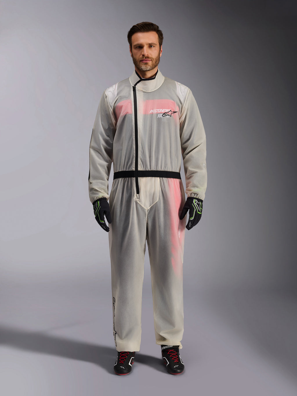 Alpinestars Kart Rain Suit, Clear, sobretraje impermeable transparente usado por un modelo, con cremallera asimétrica negra y logotipo en el pecho, mono de carreras rojo y blanco visible debajo, combinado con guantes negros y verde neón y botas de carreras negras.