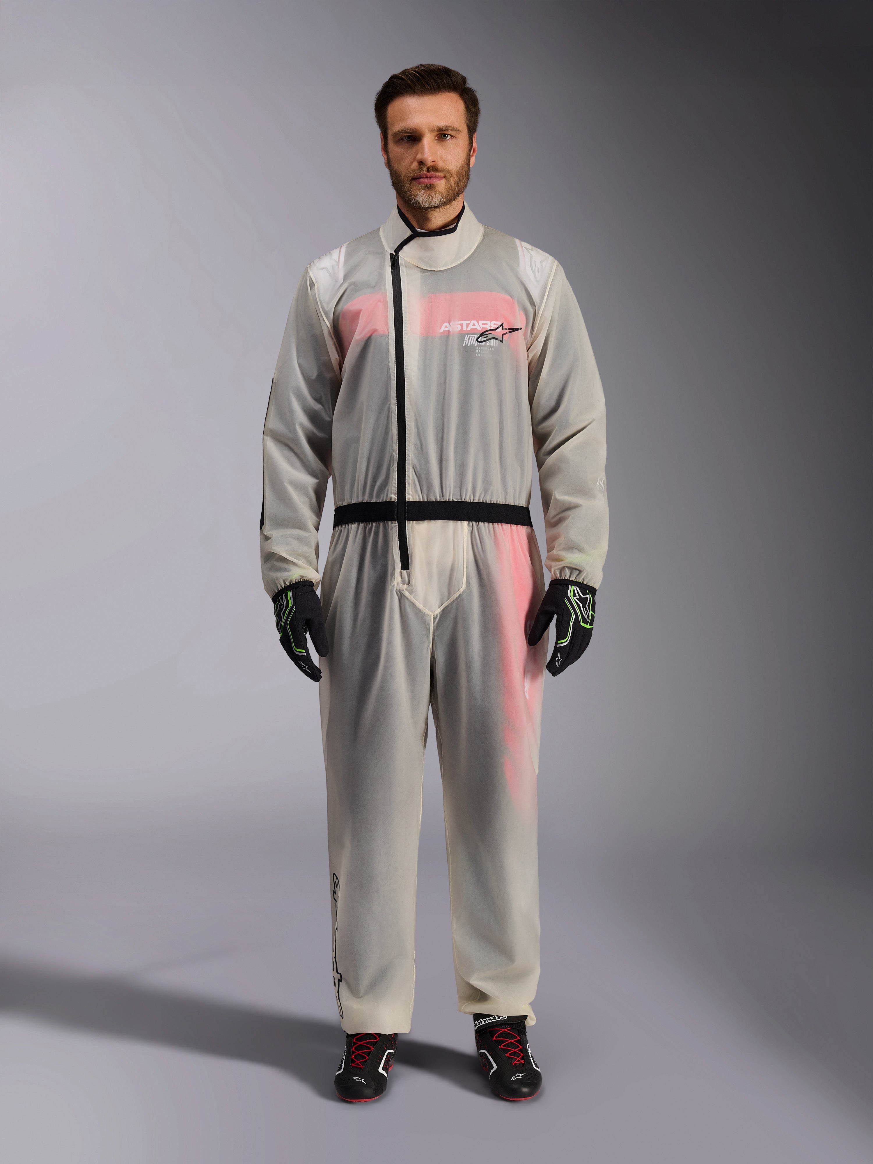 Alpinestars Kart Rain Suit, Clear, sobretraje impermeable transparente usado por un modelo, con cremallera asimétrica negra y logotipo en el pecho, mono de carreras rojo y blanco visible debajo, combinado con guantes negros y verde neón y botas de carreras negras.