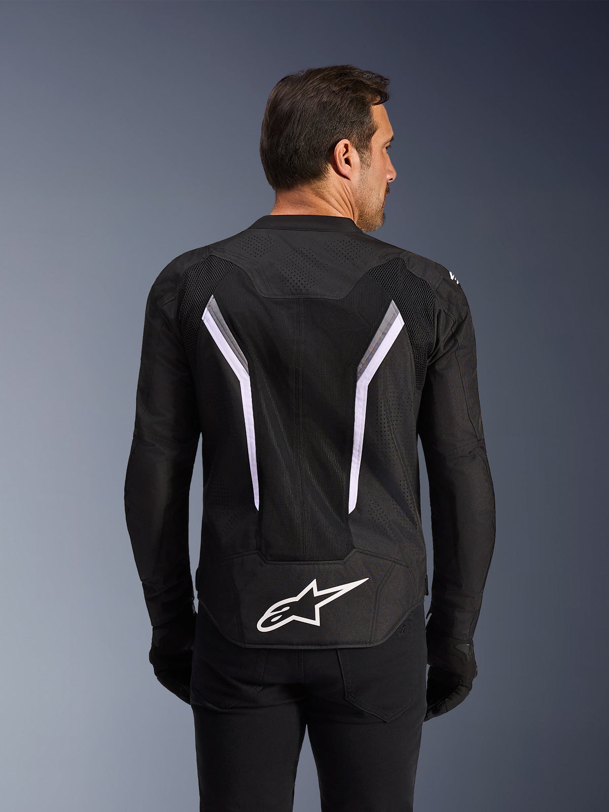 Alpinestars AST V3 Air Jacket, chaqueta de moto, negra con detalles en blanco y gris oscuro, vista trasera que muestra extensos paneles de ventilación de malla, grandes franjas blancas en forma de V y el logotipo blanco de Alpinestars en la parte inferior de la espalda, usada con guantes y pantalones negros
