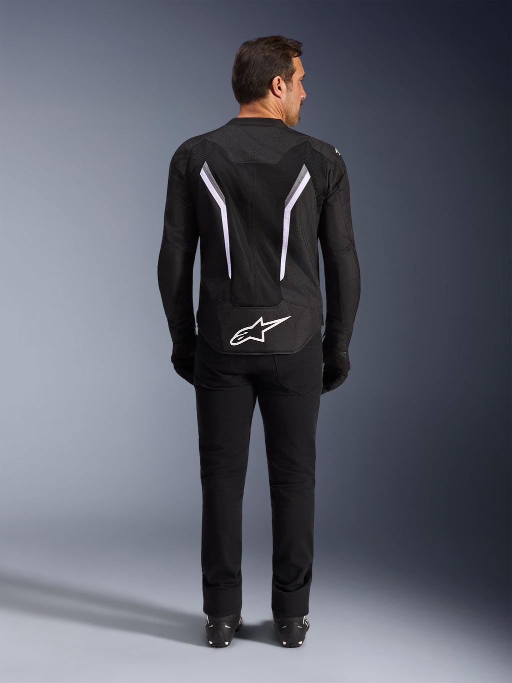 Chaqueta Alpinestars AST V3 Air, vista trasera, negro y gris oscuro con detalles blancos, con un gran logotipo Astar blanco en la parte inferior trasera, franjas reflectantes en la espalda y construcción de tejido de malla, combinada con pantalones y botas negras para un look deportivo