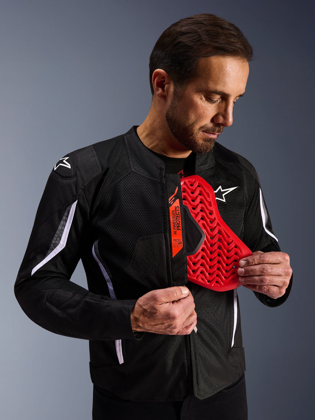 Chaqueta Alpinestars AST V3 Air en color negro, hombre mostrando una inserción de protector de pecho roja brillante con patrón de panal, destacando la compatibilidad Tech-Air Ready y la armadura interna integrada para seguridad en motocicleta.
