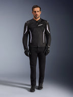 Modelo masculino vistiendo la Chaqueta Alpinestars AST V3 Air en color negro con detalles blancos, con paneles de ventilación de malla y un ajuste deportivo, combinada con pantalones técnicos de conducción, guantes y botas negros, vista frontal, equipamiento técnico de motocicleta para conducción en climas cálidos.