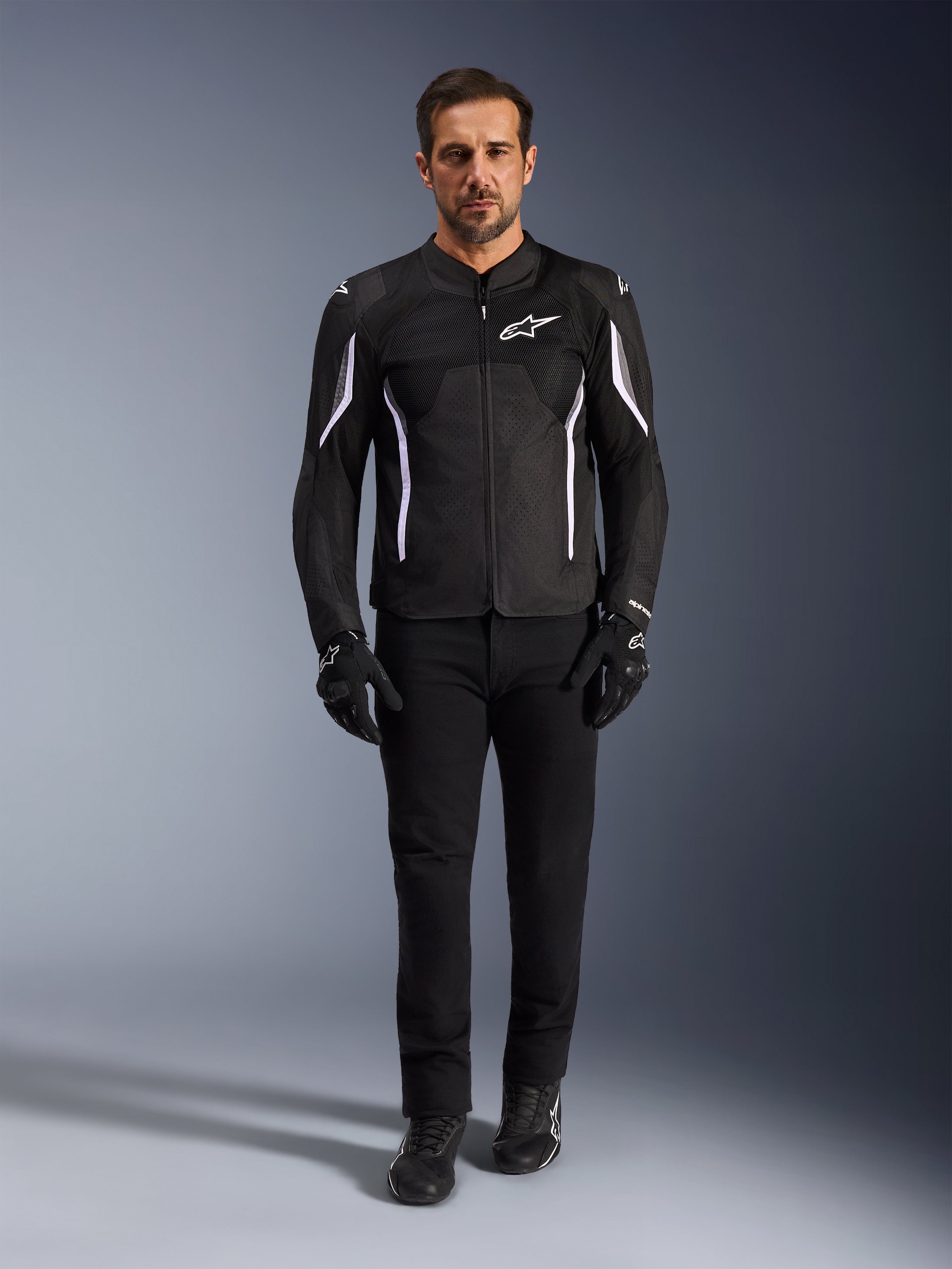 Modelo masculino vistiendo la Chaqueta Alpinestars AST V3 Air en color negro con detalles blancos, con paneles de ventilación de malla y un ajuste deportivo, combinada con pantalones técnicos de conducción, guantes y botas negros, vista frontal, equipamiento técnico de motocicleta para conducción en climas cálidos.