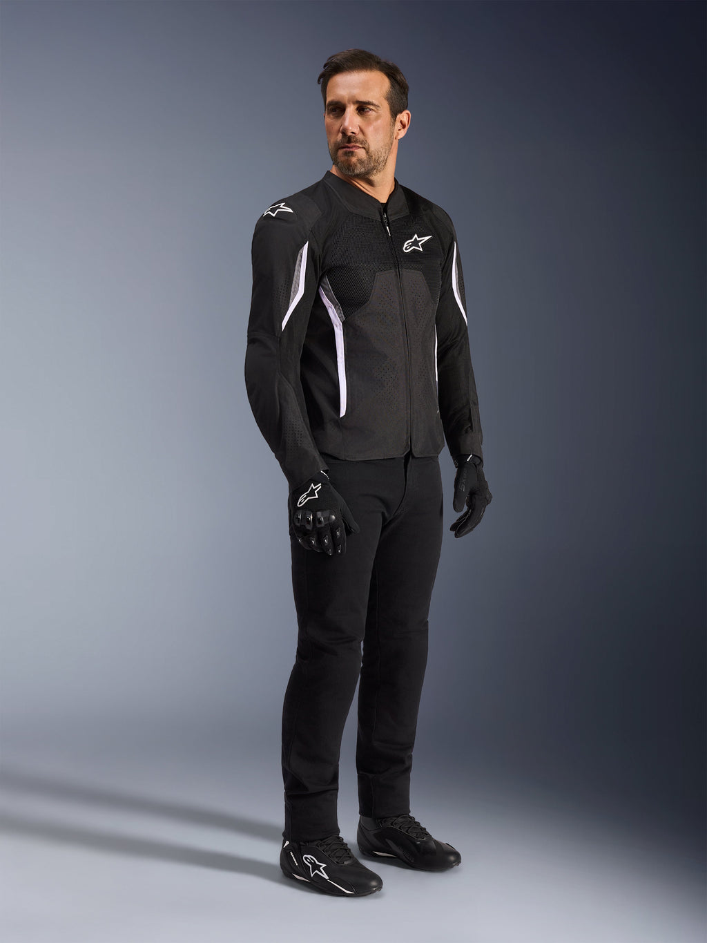 Chaqueta Alpinestars AST V3 Air, negra con detalles y logotipos en blanco, chaqueta ligera de motociclismo con ventilación de malla, usada por un hombre con pantalones, guantes y calzado de moto negros, equipamiento técnico de verano para conducción deportiva y urbana.