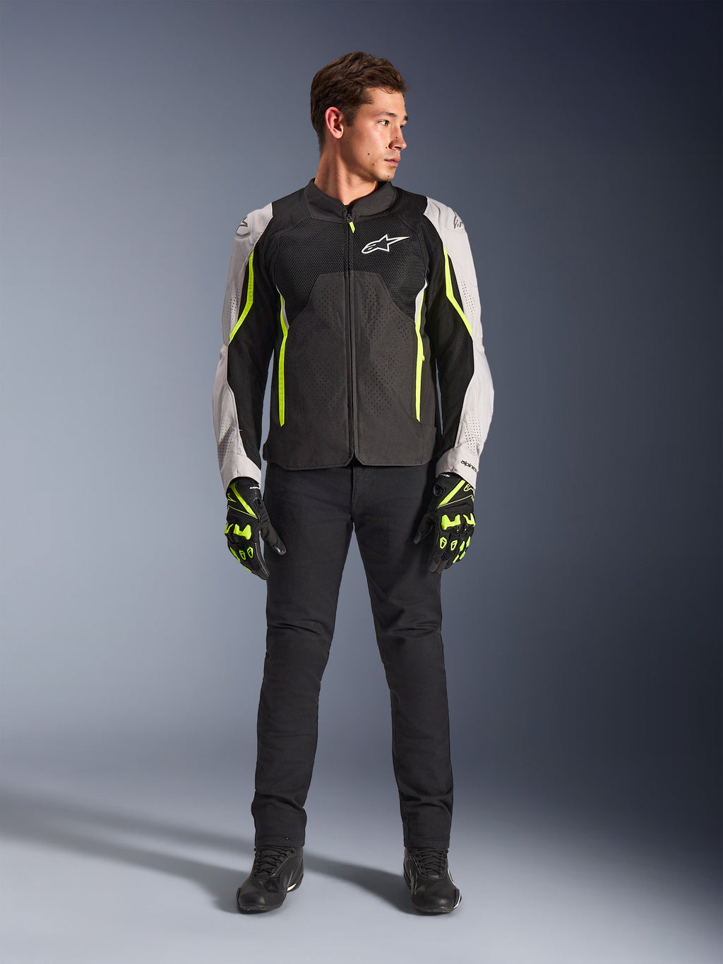 Modelo masculino con la chaqueta Alpinestars AST V3 Air en negro, plata y amarillo flúor, con paneles de ventilación de malla y detalles fluorescentes, combinada con pantalones de conducción negros y guantes de moto a juego, equipo de alto rendimiento para conducción deportiva.