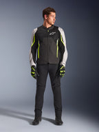 Modelo masculino con la chaqueta Alpinestars AST V3 Air en negro, plata y amarillo flúor, con paneles de ventilación de malla y detalles fluorescentes, combinada con pantalones de conducción negros y guantes de moto a juego, equipo de alto rendimiento para conducción deportiva.