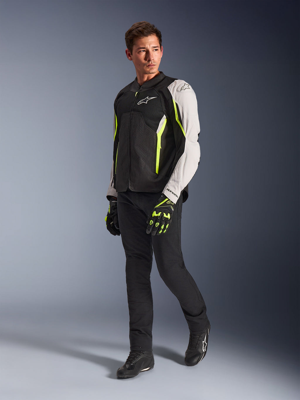 Alpinestars AST V3 Air Jacket, chaqueta de moto textil ligera en negro, plata y amarillo flúor, vestida por un modelo con pantalones negros y guantes a juego, con paneles de malla y perforaciones láser para conducción deportiva en climas cálidos.