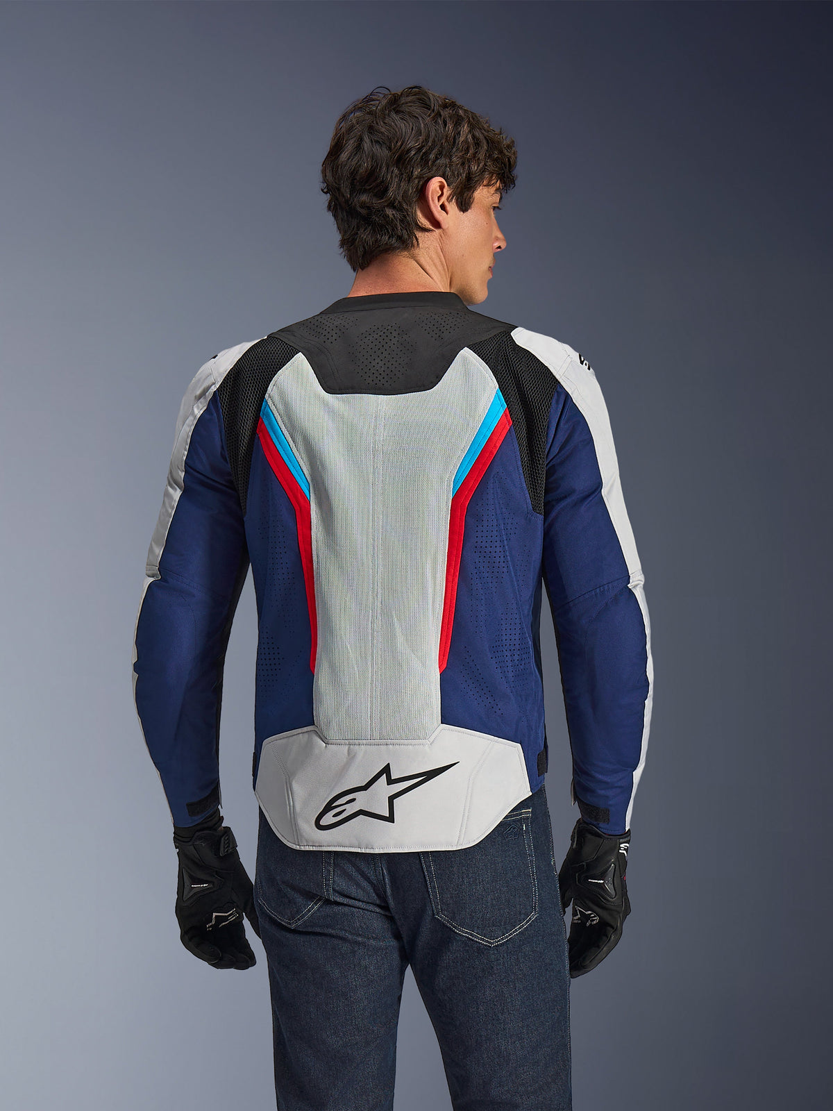 Chaqueta Alpinestars AST V3 Air, chaqueta de moto, Silver Deep Blue Bright Red, vista trasera usada por un piloto, con paneles de malla técnica y perforaciones láser para ventilación, logotipo de estrella negra en la parte inferior de la espalda, diseño deportivo optimizado para climas cálidos