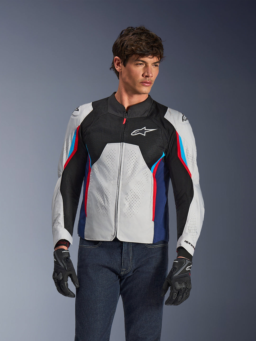 Chaqueta Alpinestars AST V3 Air en Silver Deep Blue Bright Red, vestida por un modelo con jeans oscuros y guantes negros, con paneles plateados perforados, ventilación de malla negra y detalles deportivos en rojo y azul.