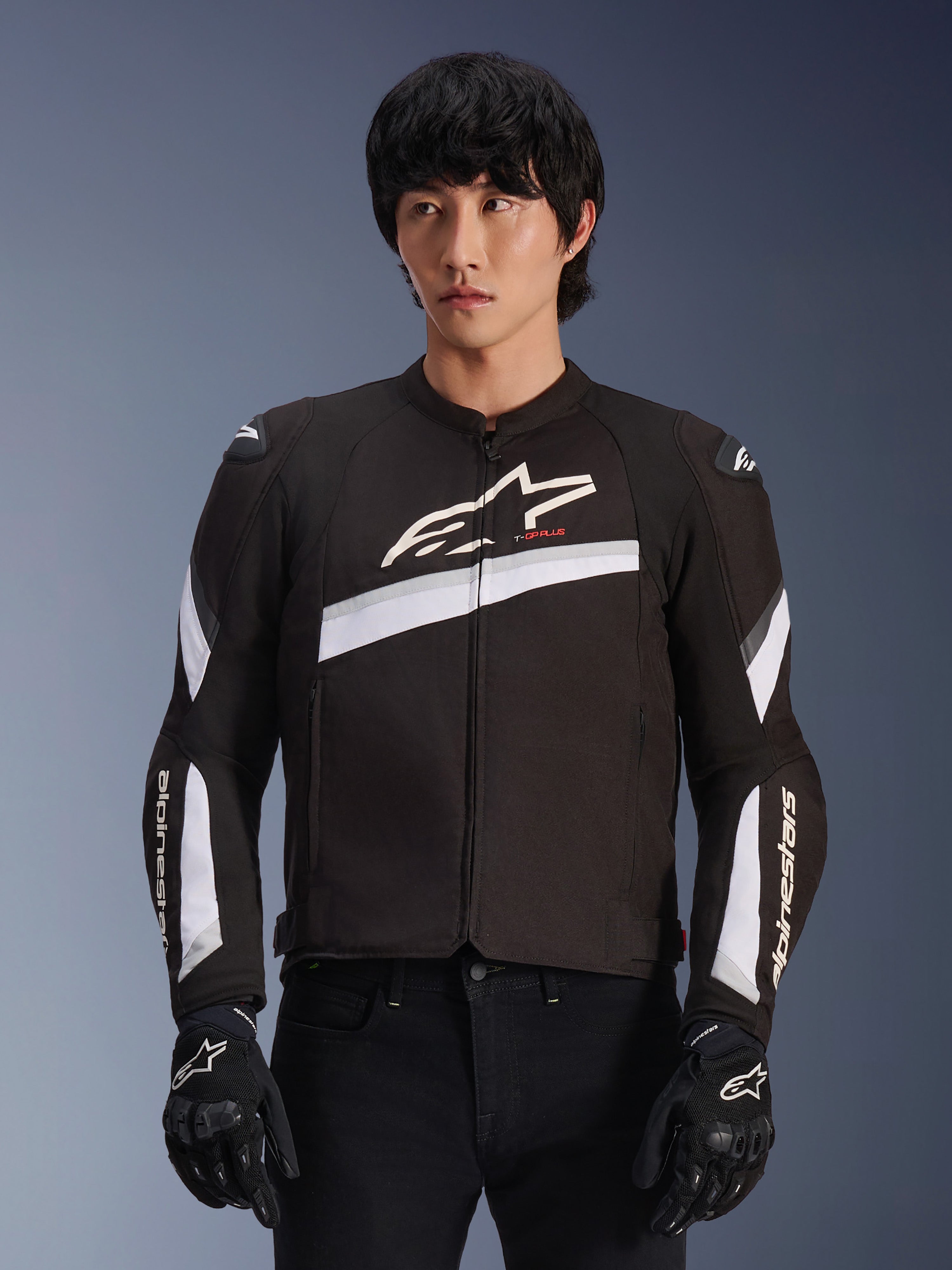 Alpinestars T-GP Plus R V4 Jacket, negro y blanco, chaqueta de moto textil, con acentos diagonales blancos y logos de la marca en el pecho y las mangas, usada por un modelo con guantes de moto negros, diseño deportivo ajustado para conducción en carretera