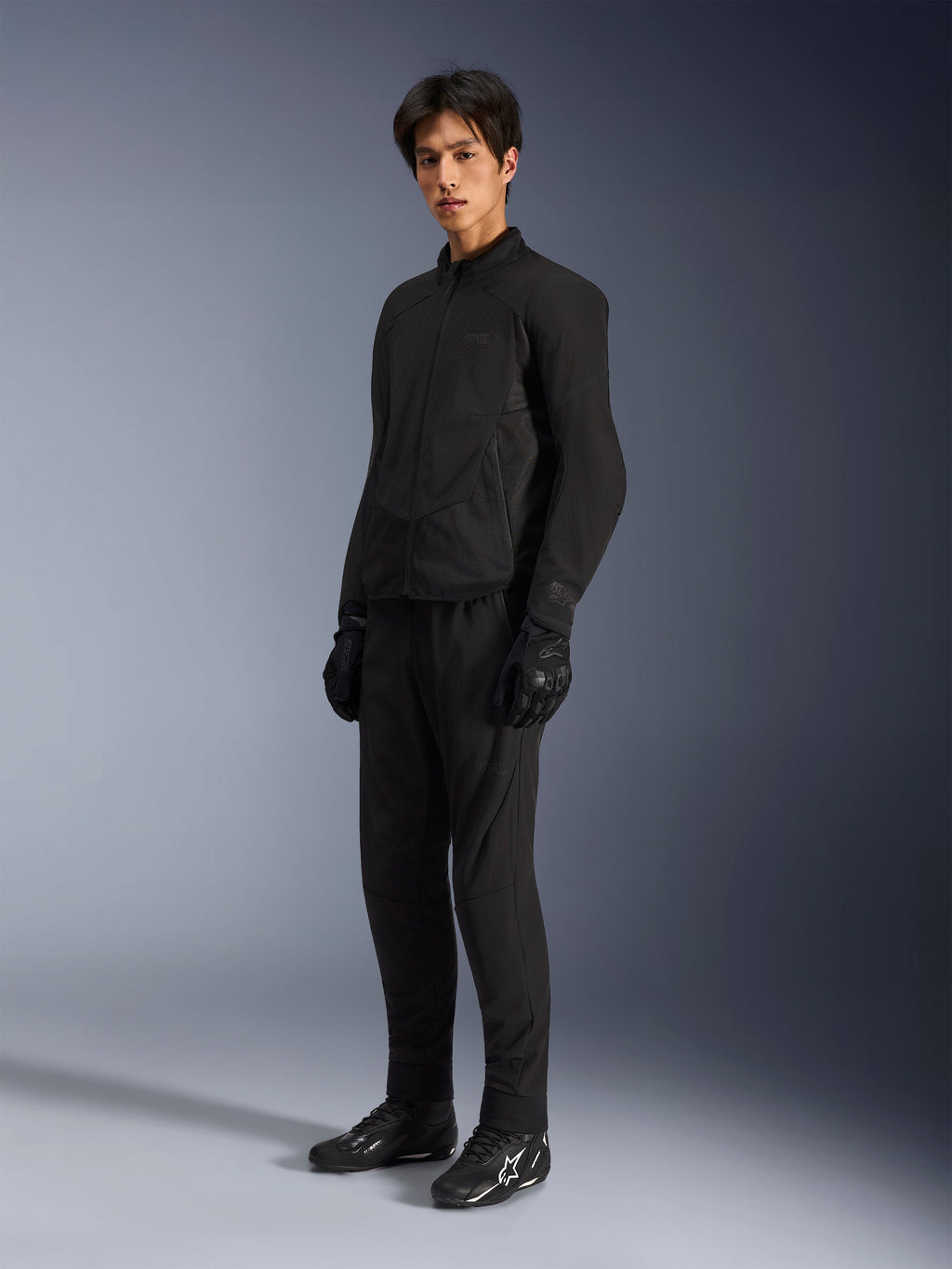Chaqueta Alpinestars Aeroshell Airflow, chaqueta de moto Black Anthracite, con ventilación perforada con láser y paneles de malla para el flujo de aire, lucida por un modelo masculino con pantalones de motorista negros a juego, guantes y botas negras con logotipos blancos, diseño de corte deportivo para rendimiento en carretera