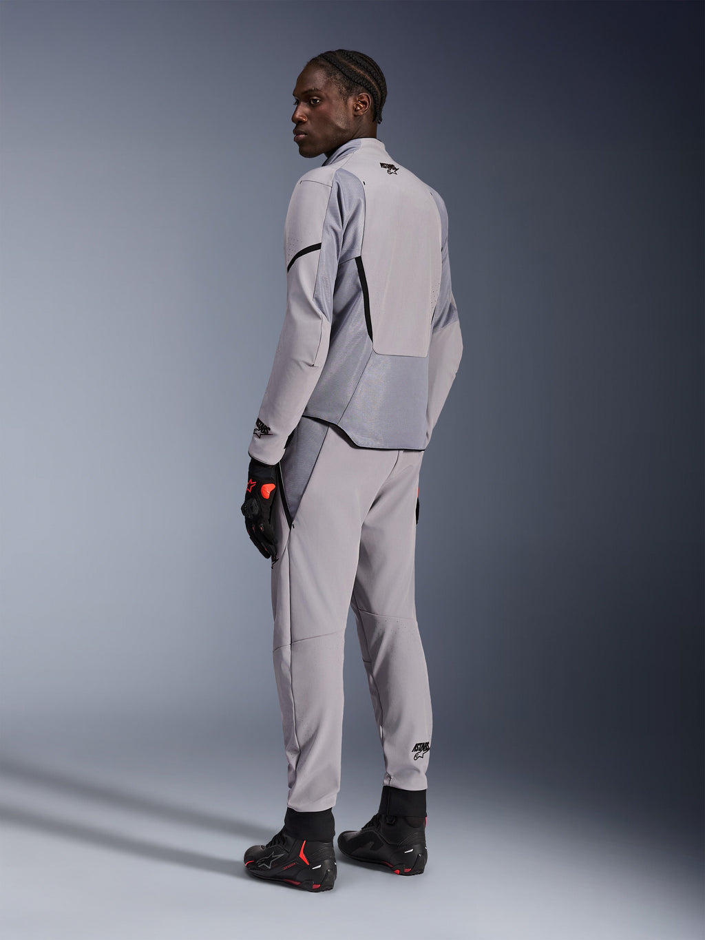 Chaqueta Alpinestars Aeroshell Airflow en color Mid Gray Anthracite, combinada con pantalones a juego y guantes de carreras negros, vista trasera que muestra los paneles perforados por láser y el ajuste deportivo, equipamiento profesional para motociclismo