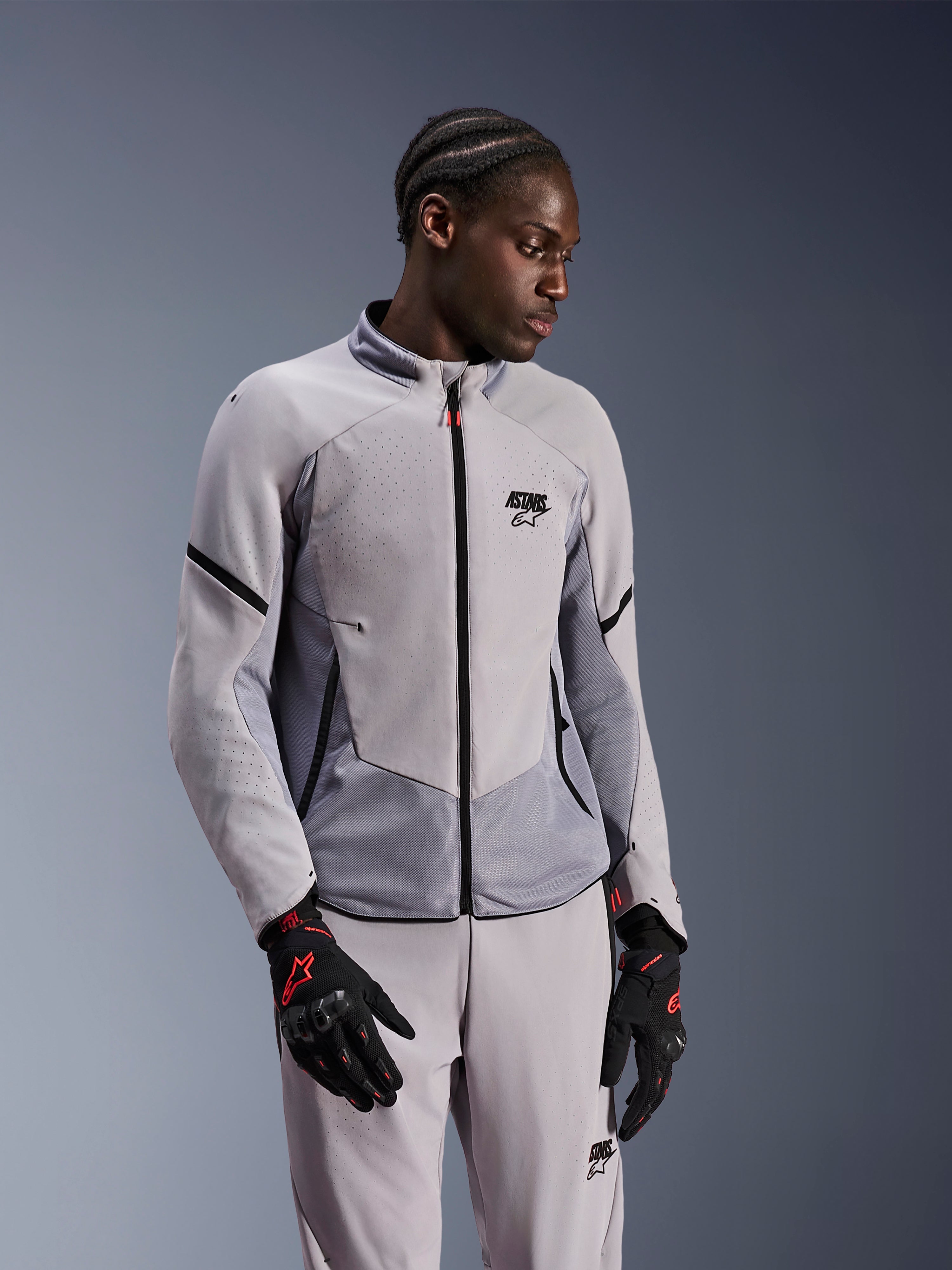 Chaqueta Alpinestars Aeroshell Airflow, Mid Gray Anthracite, usada por un modelo con pantalones textiles a juego y guantes negros con detalles rojos, con zonas perforadas por láser y paneles de malla para ventilación, diseño estilizado de ajuste deportivo para la conducción de motocicletas.