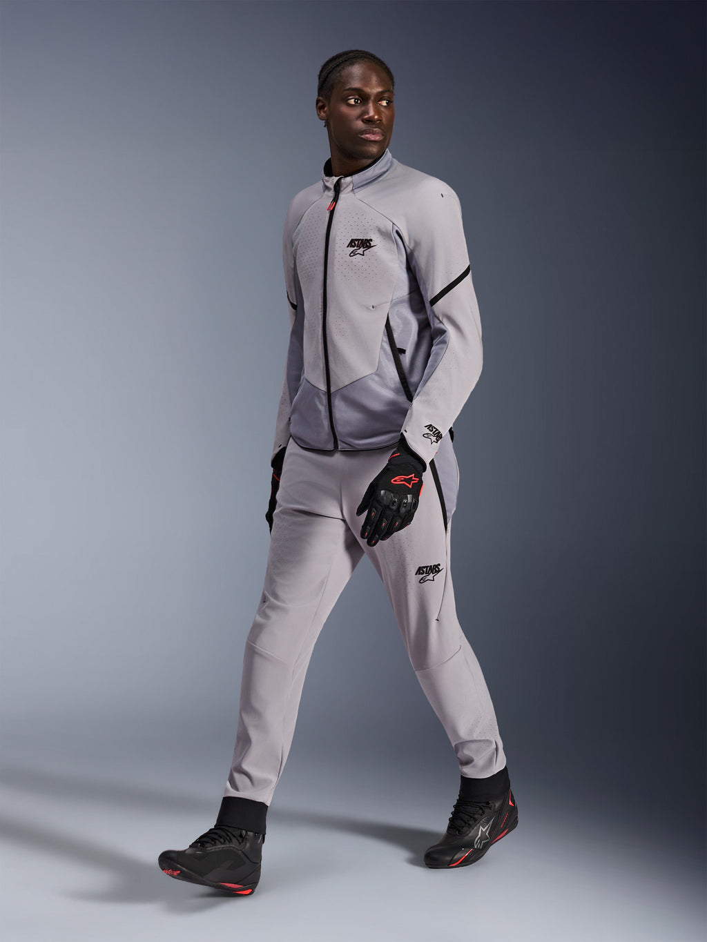 Chaqueta Alpinestars Aeroshell Airflow en color Mid Gray Anthracite, lucida por un modelo masculino con pantalones de pilotaje grises a juego y guantes negros, con ventilación en el pecho perforada por láser y un elegante diseño de corte deportivo.