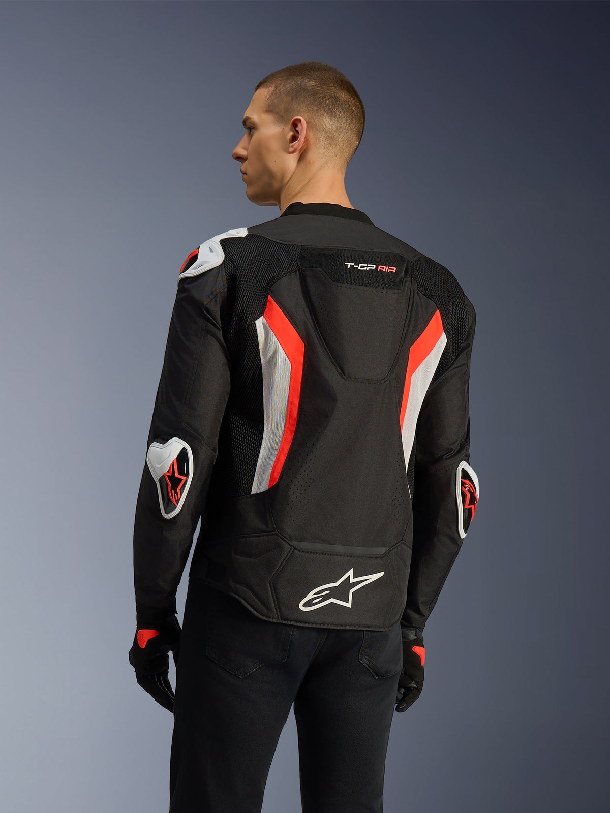Chaqueta Alpinestars T-GP Air en Black Red Fluo, vista trasera de tres cuartos en un modelo, destacando la joroba aerodinámica, protecciones externas Racing GP DFS en hombros y codos, paneles de malla perforados por láser y detalles en blanco y rojo flúor, chaqueta de moto textil de corte deportivo.