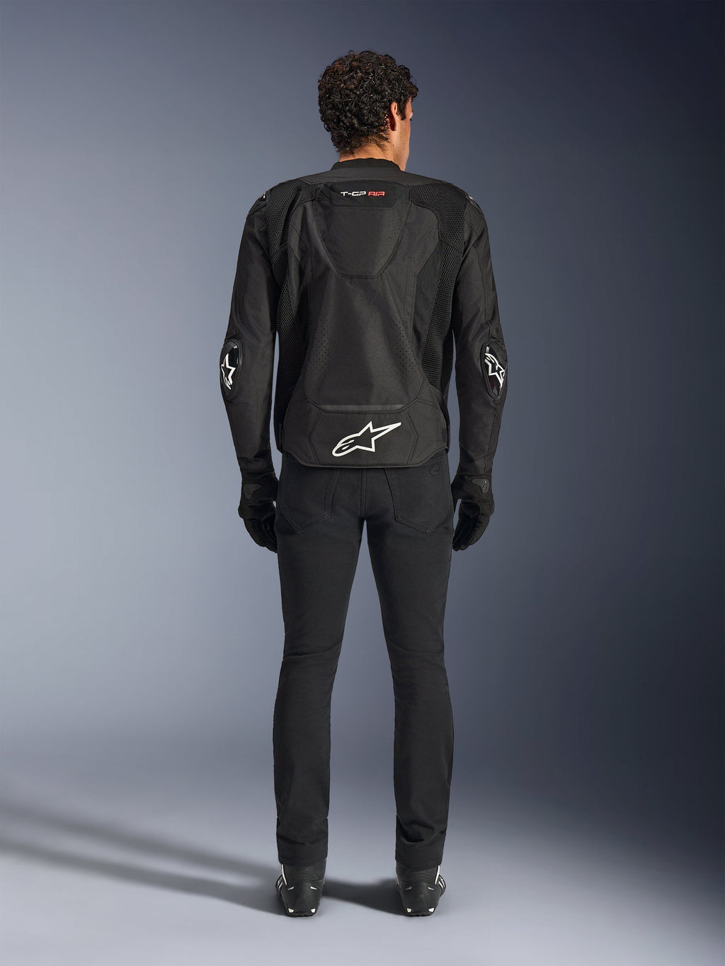 Chaqueta Alpinestars T-GP Air, negra, vista trasera de un hombre con la chaqueta textil de motociclismo, con paneles de ventilación de malla, joroba aerodinámica y logotipos blancos de Alpinestars en la parte inferior de la espalda y los codos, combinada con pantalones y guantes de conducción negros