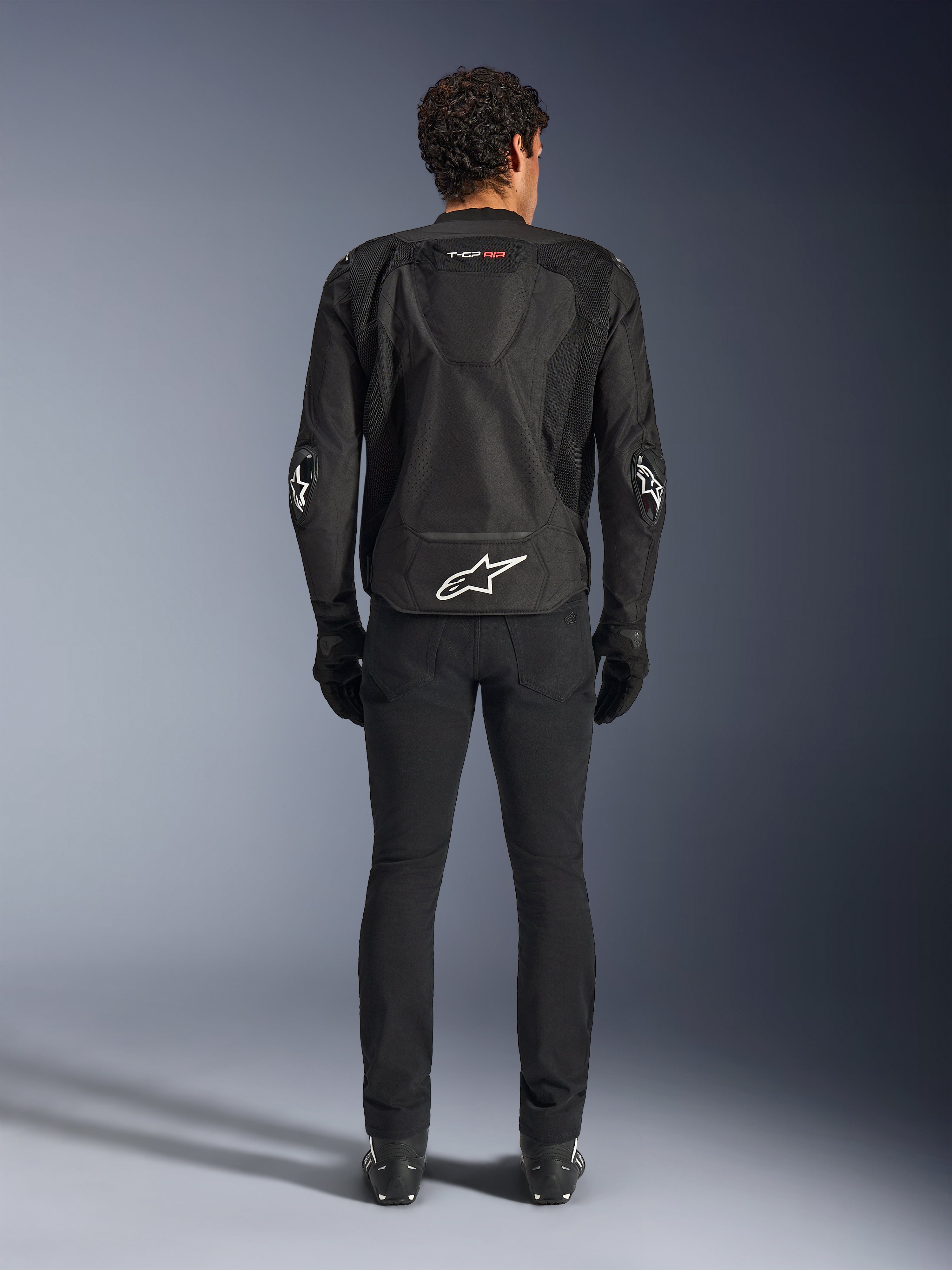 Chaqueta Alpinestars T-GP Air, negra, vista trasera de un hombre con la chaqueta textil de motociclismo, con paneles de ventilación de malla, joroba aerodinámica y logotipos blancos de Alpinestars en la parte inferior de la espalda y los codos, combinada con pantalones y guantes de conducción negros
