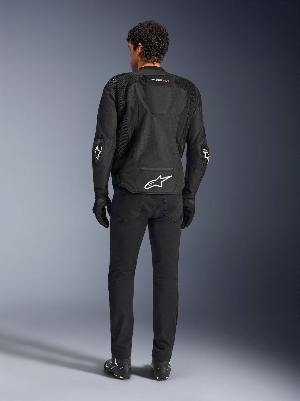 Chaqueta Alpinestars T-GP Air, negra, vista posterior en modelo, que muestra los paneles de ventilación de malla 3D y la joroba aerodinámica, con el logotipo de la estrella blanca en la parte inferior de la espalda y los codos, usada con pantalones y guantes de moto negros, indumentaria de conducción deportiva.