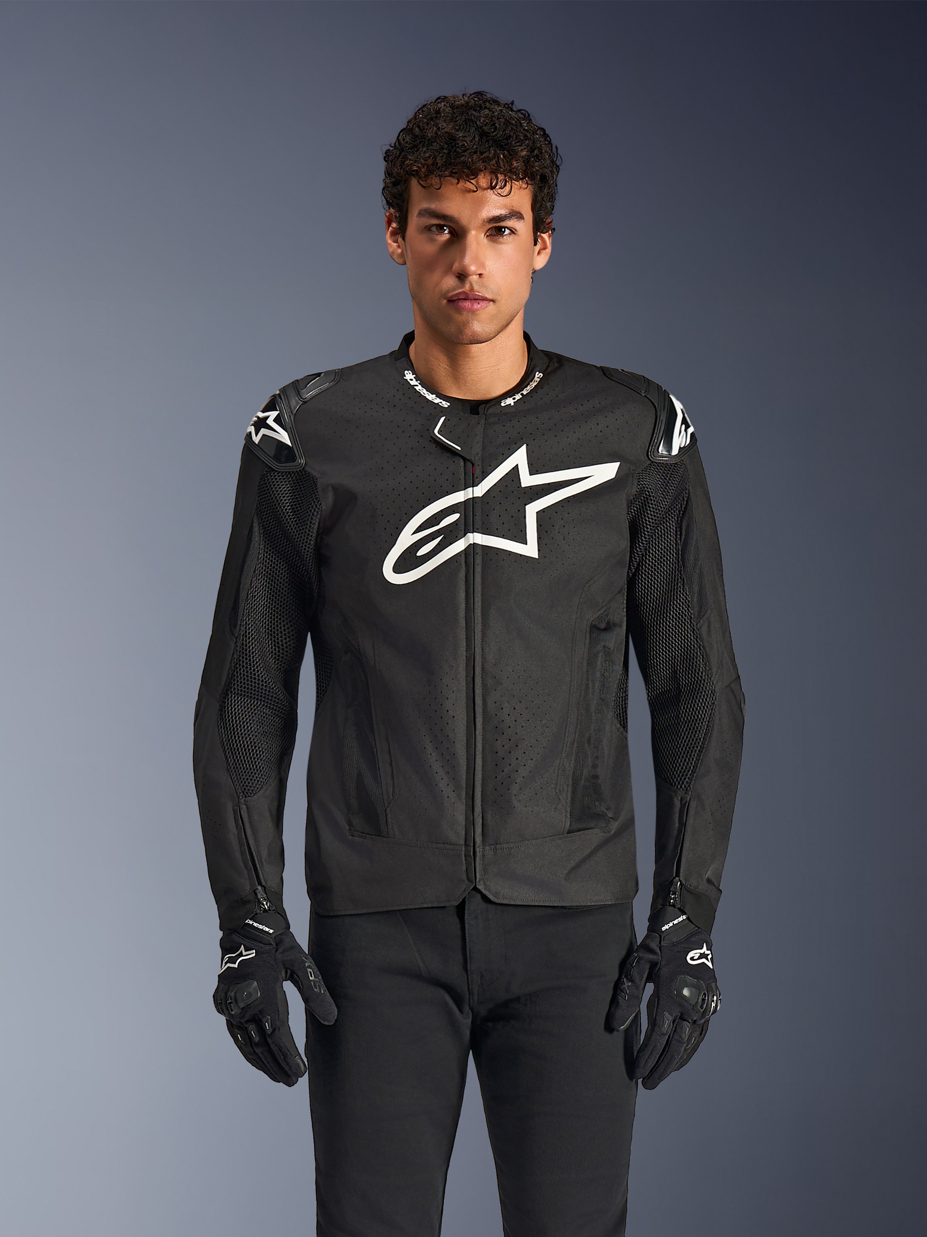 Alpinestars T-GP Air Jacket, chaqueta de moto textil negra con un gran logotipo de estrella blanca en el pecho, paneles de ventilación perforados y malla 3D, deslizadores externos en los hombros, usada con guantes negros y blancos, diseño deportivo para conducción en carretera en climas cálidos