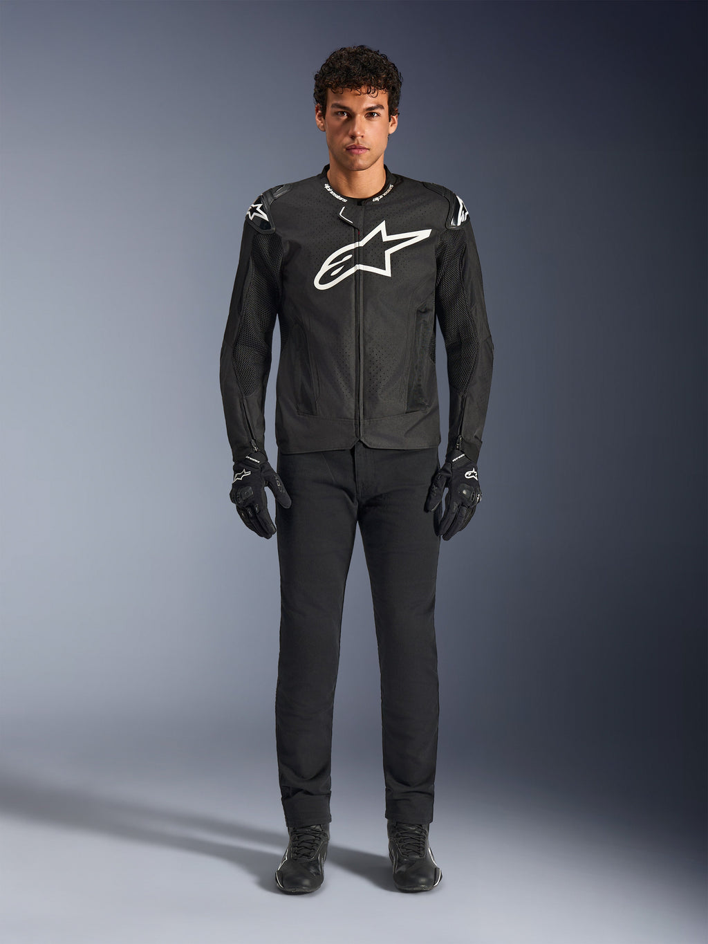 Chaqueta Alpinestars T-GP Air, chaqueta de moto textil negra con un gran logotipo Astars blanco en el pecho, usada por un modelo con pantalones de conducción y guantes negros, con paneles perforados y ventilación de malla para conducción deportiva