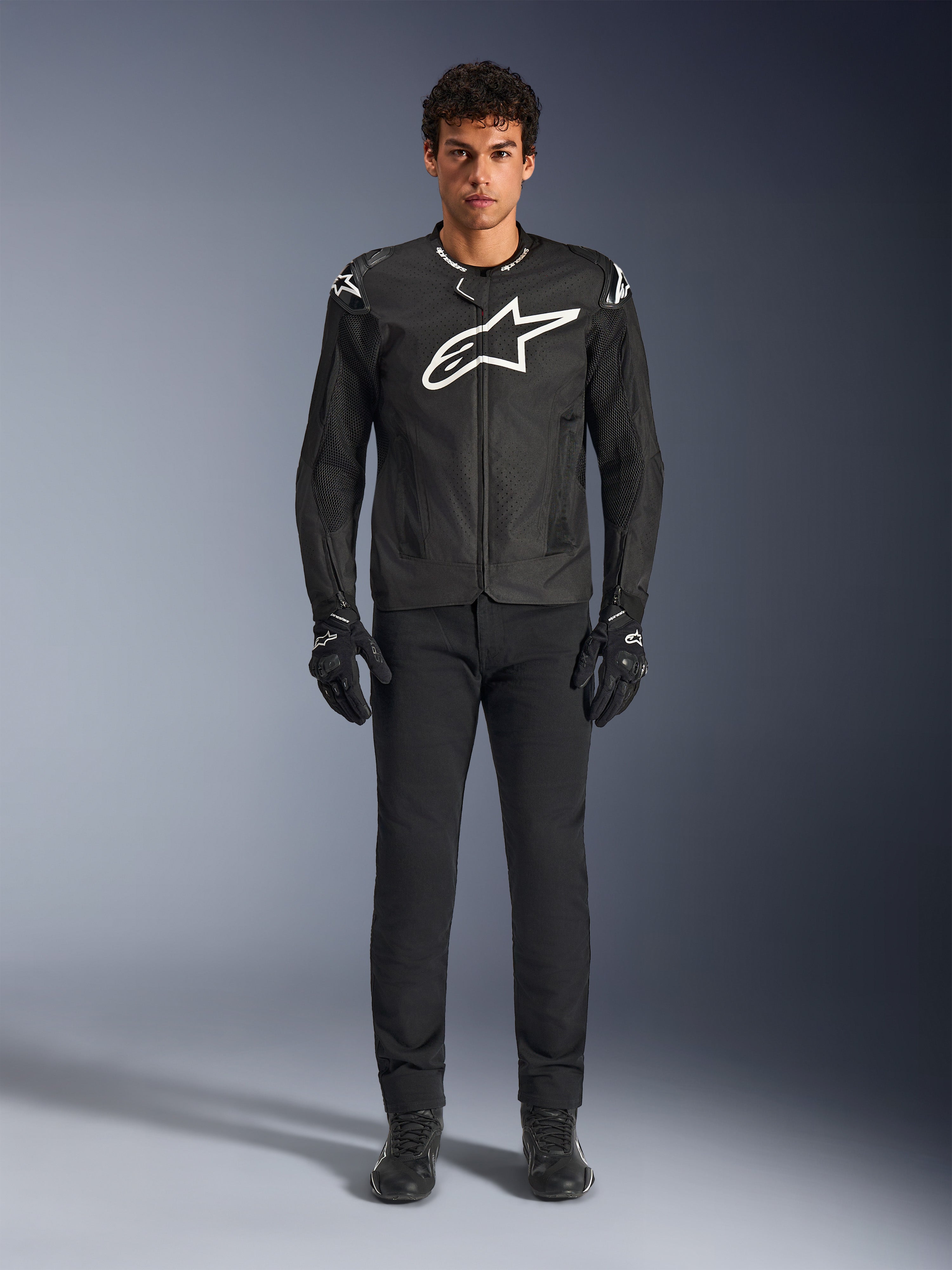 Chaqueta Alpinestars T-GP Air, chaqueta de moto textil negra con un gran logotipo Astars blanco en el pecho, usada por un modelo con pantalones de conducción y guantes negros, con paneles perforados y ventilación de malla para conducción deportiva
