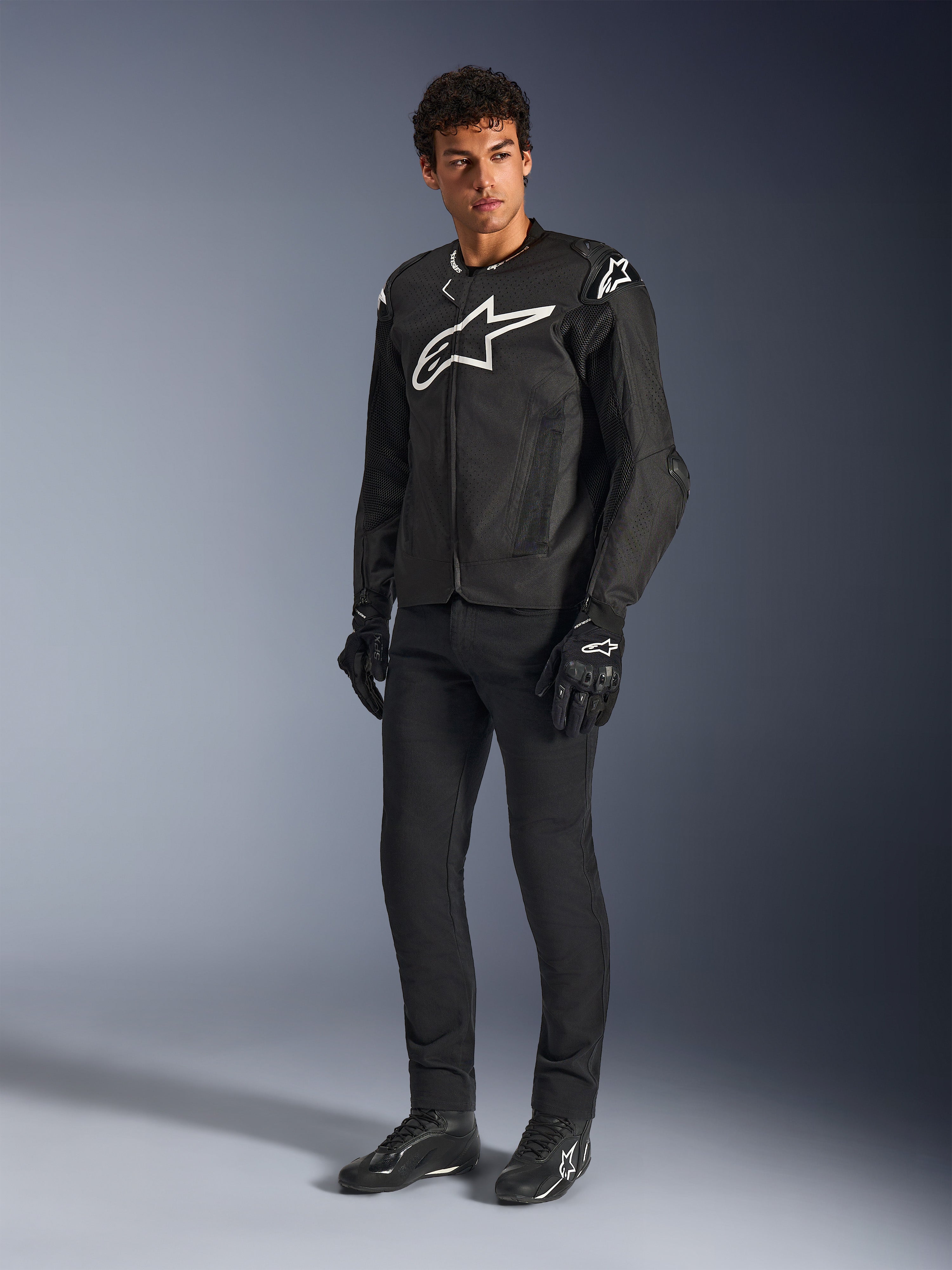Chaqueta Alpinestars T-GP Air, chaqueta de motociclismo textil en color negro, vestida por un modelo con pantalones y guantes negros a juego, con un destacado logotipo de Alpinestars en blanco en el pecho y los hombros, paneles de malla ventilados y un ajuste deportivo derivado de las carreras