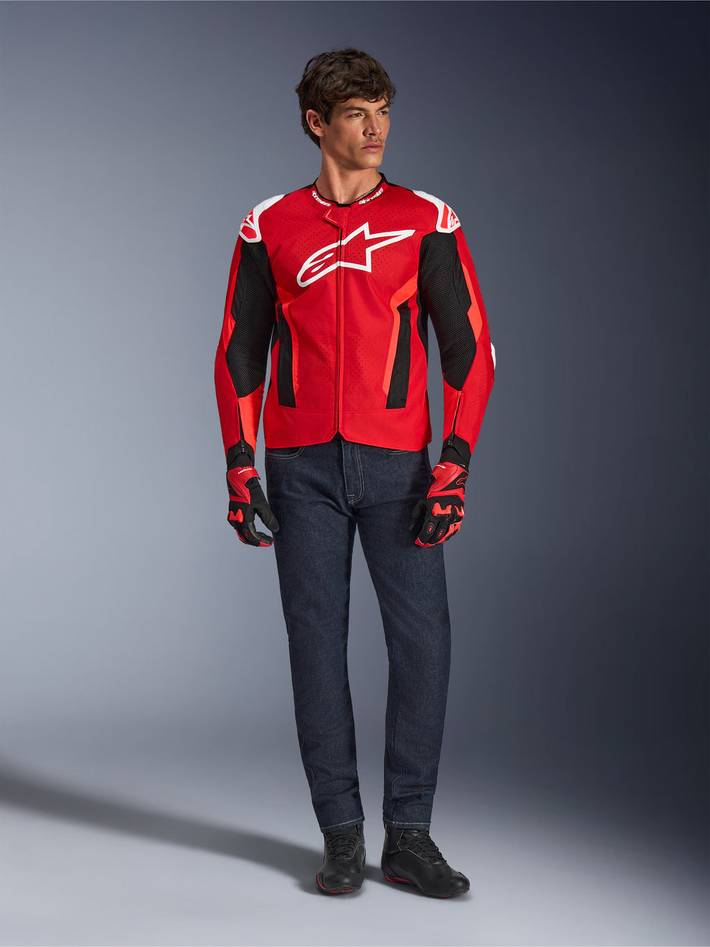 Chaqueta Alpinestars T-GP Air, rojo brillante, rojo flúor y negro, modelo masculino con equipamiento de moto completo, vaqueros azul oscuro y guantes a juego, chaqueta textil deportiva con paneles de malla perforada y logo Astars blanco, foto de estudio.