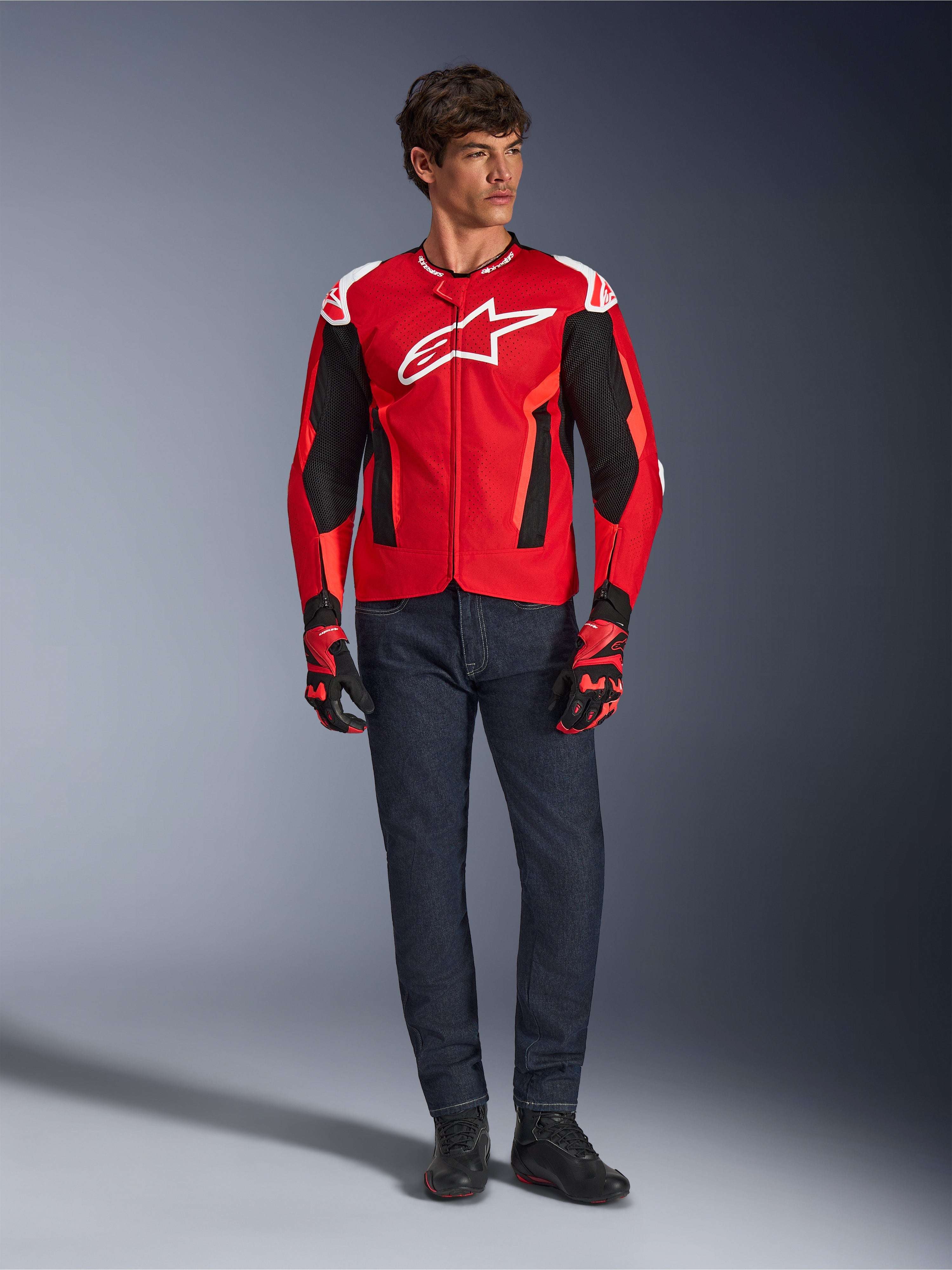 Chaqueta Alpinestars T-GP Air, rojo brillante, rojo flúor y negro, modelo masculino con equipamiento de moto completo, vaqueros azul oscuro y guantes a juego, chaqueta textil deportiva con paneles de malla perforada y logo Astars blanco, foto de estudio.
